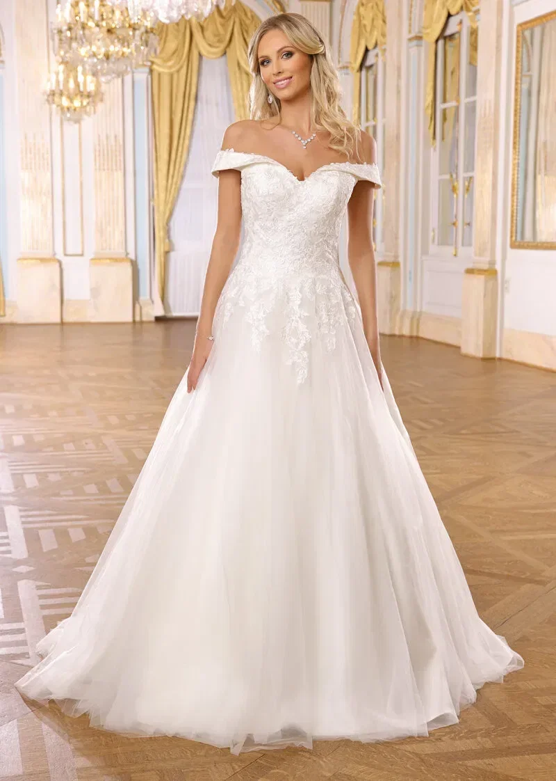Braut in einem schulterfreien, spitzenverzierten Hochzeitskleid, die in einem eleganten, goldverzierten Ballsaal posiert.