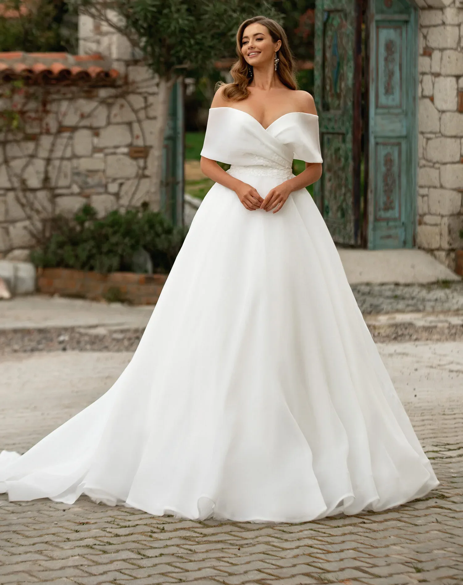 Braut in einem schulterfreien weißen Brautkleid mit voluminösem Rock, posierend vor einem rustikalen Gebäude im Freien.
