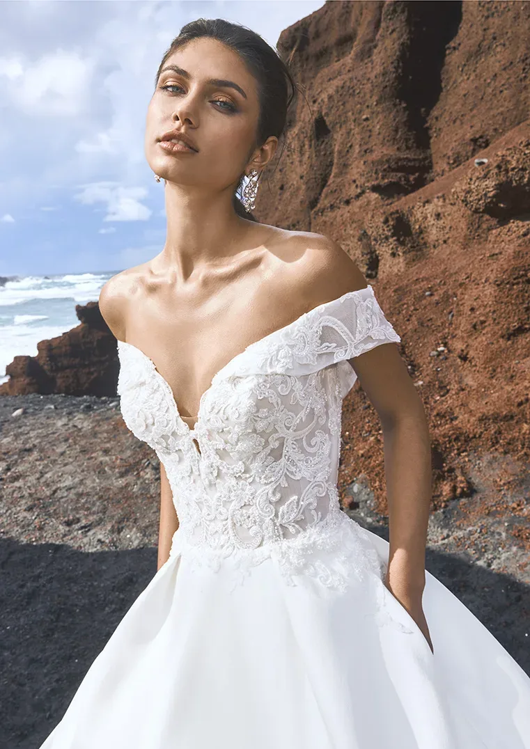 Braut in einem schulterfreien Spitzenkleid steht vor einer felsigen Küste mit Blick aufs Meer, elegant posierend unter blauem Himmel.