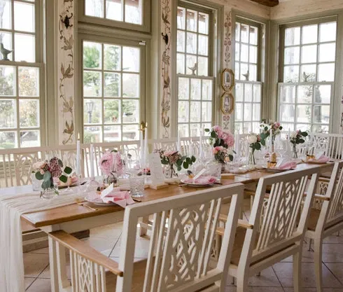 Elegant dekorierter Hochzeitstisch in einem lichtdurchfluteten Raum mit großen Fenstern, blumigen Akzenten und weiß lackierten Holzstühlen.