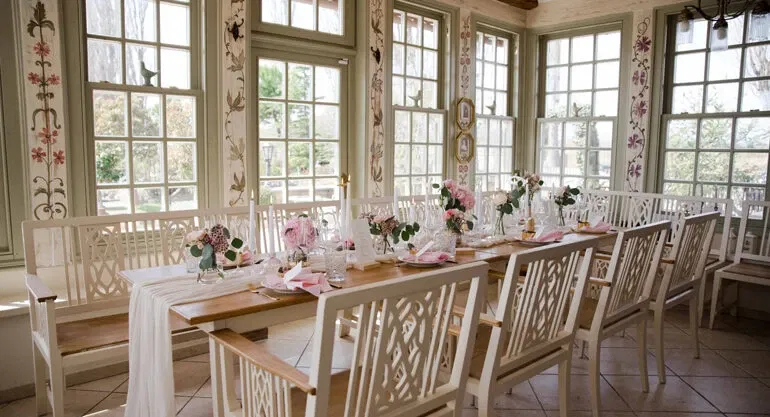 Elegant dekorierter Hochzeitstisch in einem lichtdurchfluteten Raum mit großen Fenstern, blumigen Akzenten und weiß lackierten Holzstühlen.