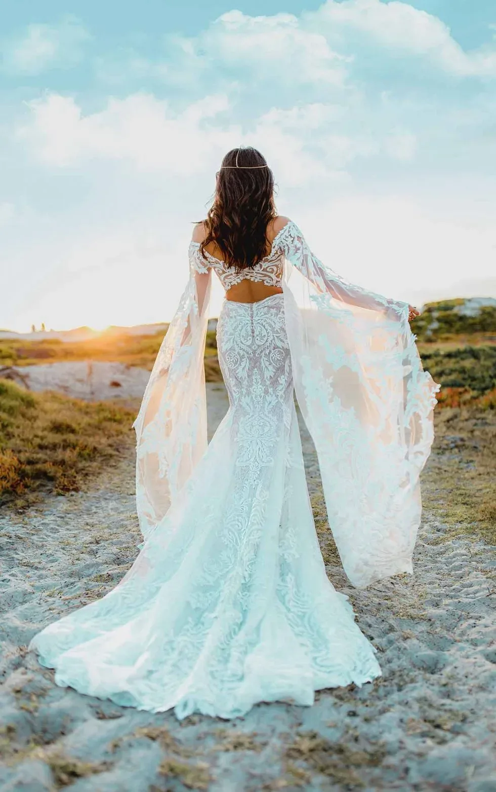 Rückansicht einer Braut in einem schulterfreien Spitzenkleid mit langen Ärmeln, die auf einem sandigen Pfad dem Sonnenuntergang entgegen geht.
