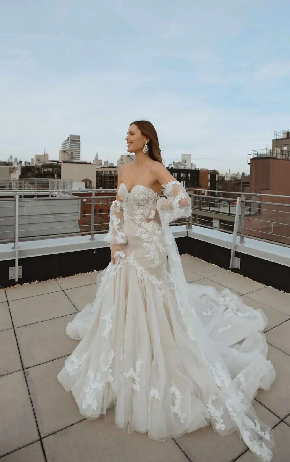 Braut in einem schulterfreien Hochzeitskleid mit floralen Applikationen und langer Schleppe, posierend auf einer Dachterrasse mit Stadtblick.