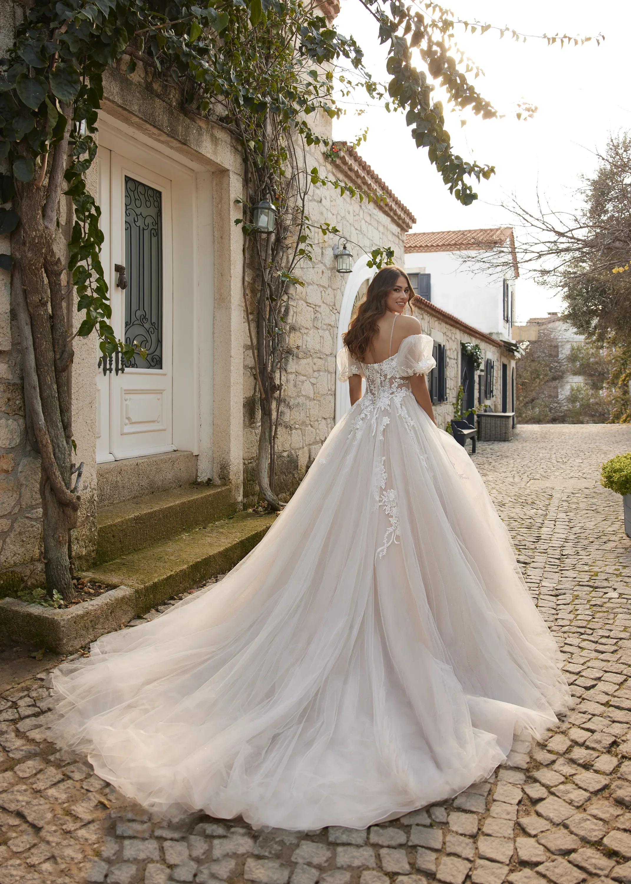 Braut in einem romantischen, spitzenverzierten Ballkleid mit langer Schleppe spaziert durch eine malerische Gasse mit Kopfsteinpflaster.