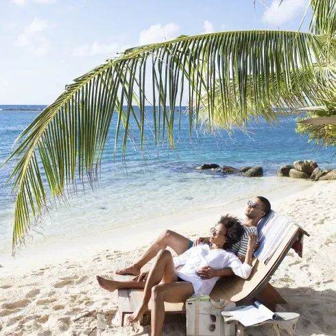 Ein Paar liegt entspannt auf einer Sonnenliege unter einer Palme am Strand. Sie genießen den Meerblick und die ruhige Atmosphäre. Im Vordergrund liegt eine Strandtasche im Sand, während das klare Wasser des Ozeans den Horizont schmückt.