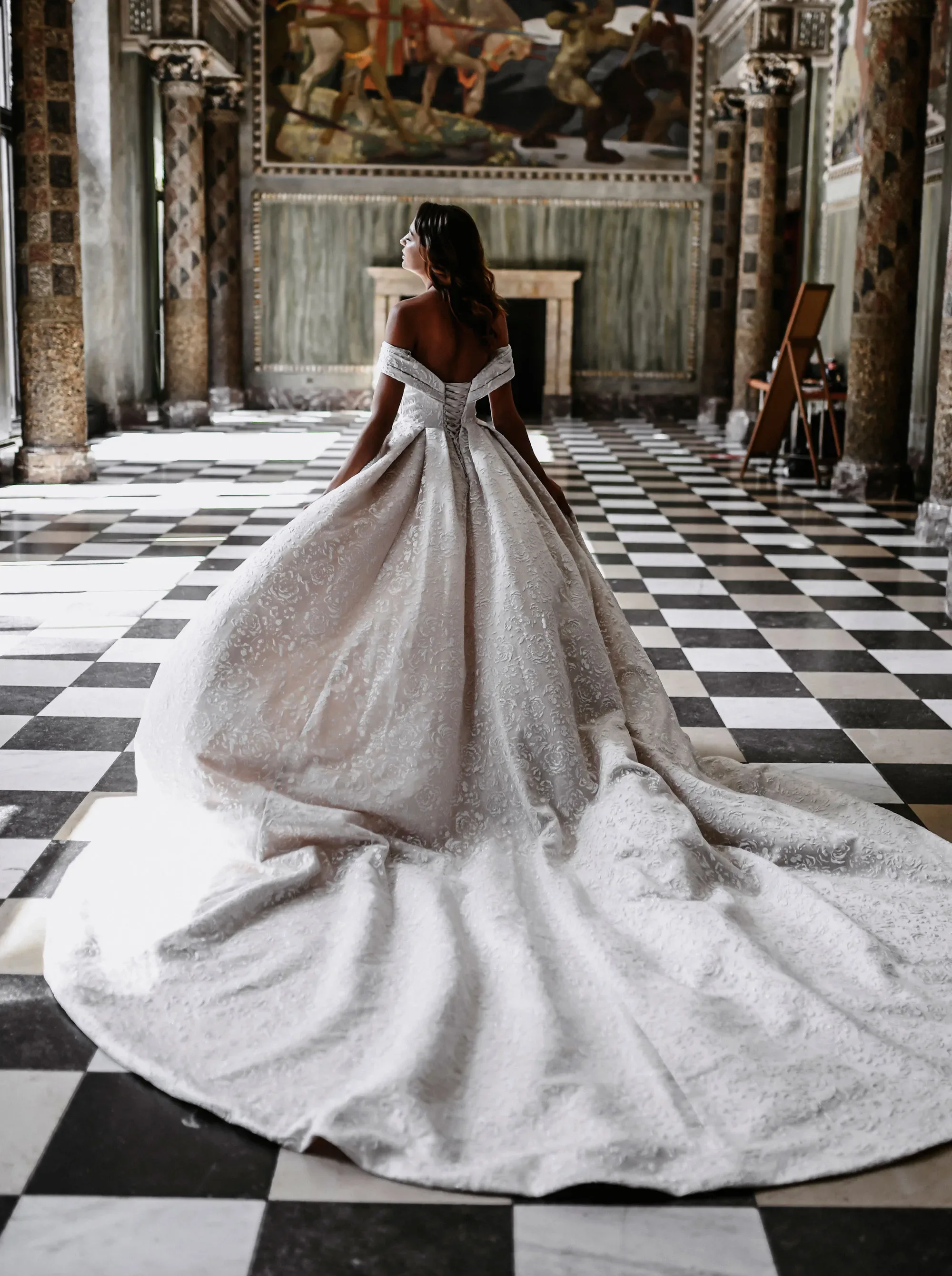 Braut in einem eleganten, schulterfreien Ballkleid mit langer Schleppe, die in einem historischen Saal mit Schachbrettboden posiert.