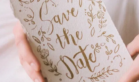 Save-the-Date-Karte in natürlichen Tönen mit verspieltem floralen Design und handgeschriebener Schrift, in der Hand gehalten.
