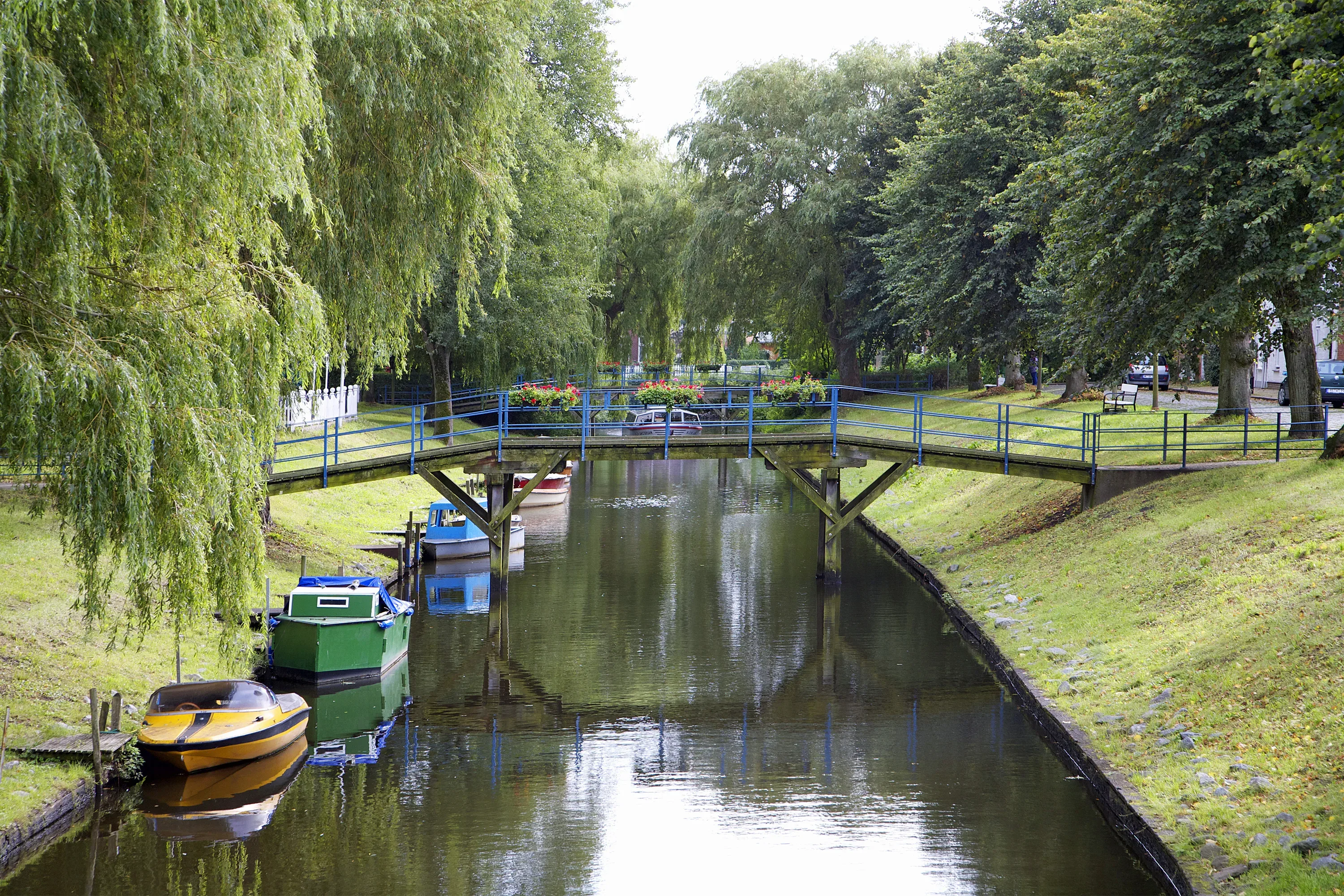 Eine idyllische Szene in Friedrichstadt mit einer kleinen blauen Brücke, die über einen von Booten gesäumten Kanal führt. Umgeben von üppigen Bäumen und Grünflächen spiegeln sich die Boote und das Geländer der Brücke im ruhigen Wasser wider.