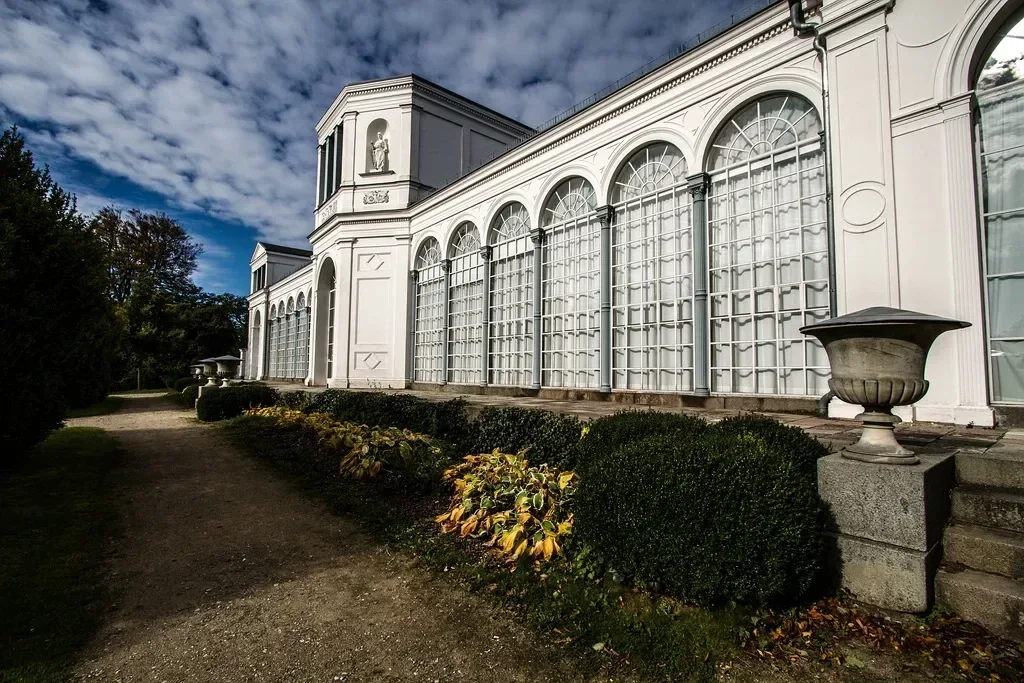 Ein weißes historisches Gebäude mit großen Bogenfenstern und dekorativen Glasfronten. Ein gepflegter Gartenweg führt entlang der Fassade, während der Himmel von Wolken bedeckt ist.