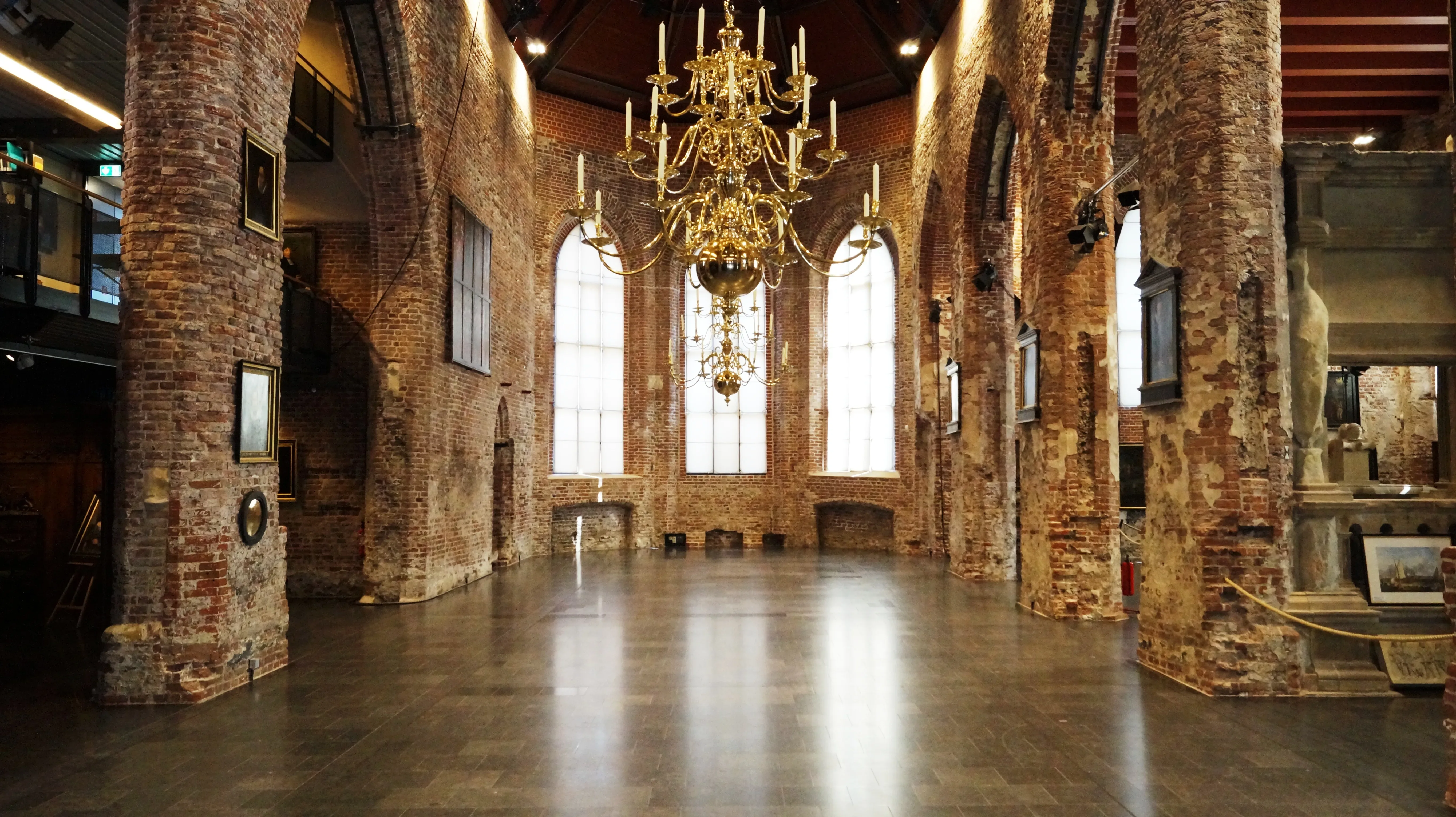 Innenansicht einer Kirchenapsis mit freigelegtem Mauerwerk aus roten Ziegeln und einem großen, goldenen Kronleuchter, der von der Decke herabhängt. Die hohen Fenster lassen sanftes Licht in den Raum fallen.