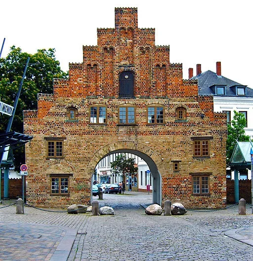 Das historische Nordertor in Flensburg, ein rotes Backsteingebäude im gotischen Stil mit einem zentralen Torbogen, durch den eine Straße führt.