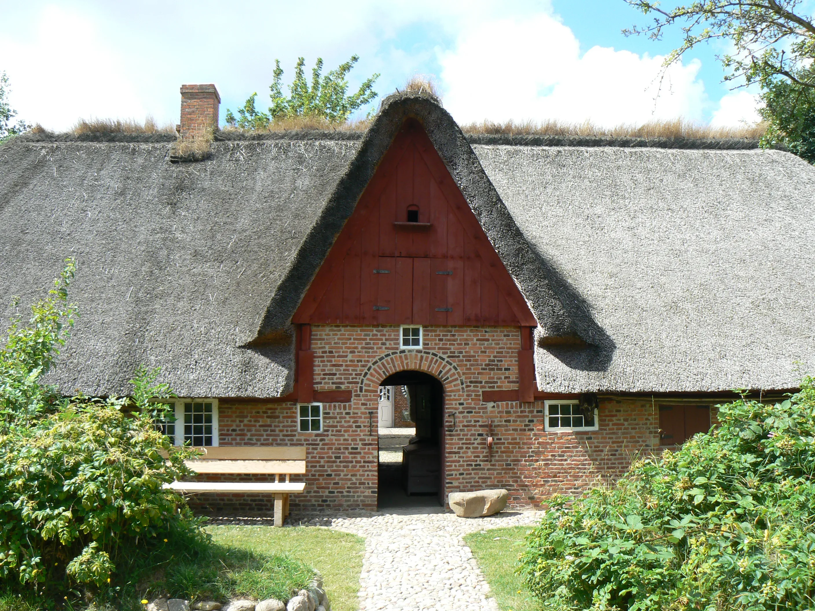 Traditionelles Reetdachhaus auf der Insel Föhr, bekannt als Haus Olesen. Es besteht aus roten Backsteinen und hat ein tiefes, mit Stroh gedecktes Dach. In der Mitte befindet sich ein Torbogen mit einer kleinen, roten Holzwand darüber. Links neben dem Gebäude steht eine Holzbank, umgeben von grünen Büschen.