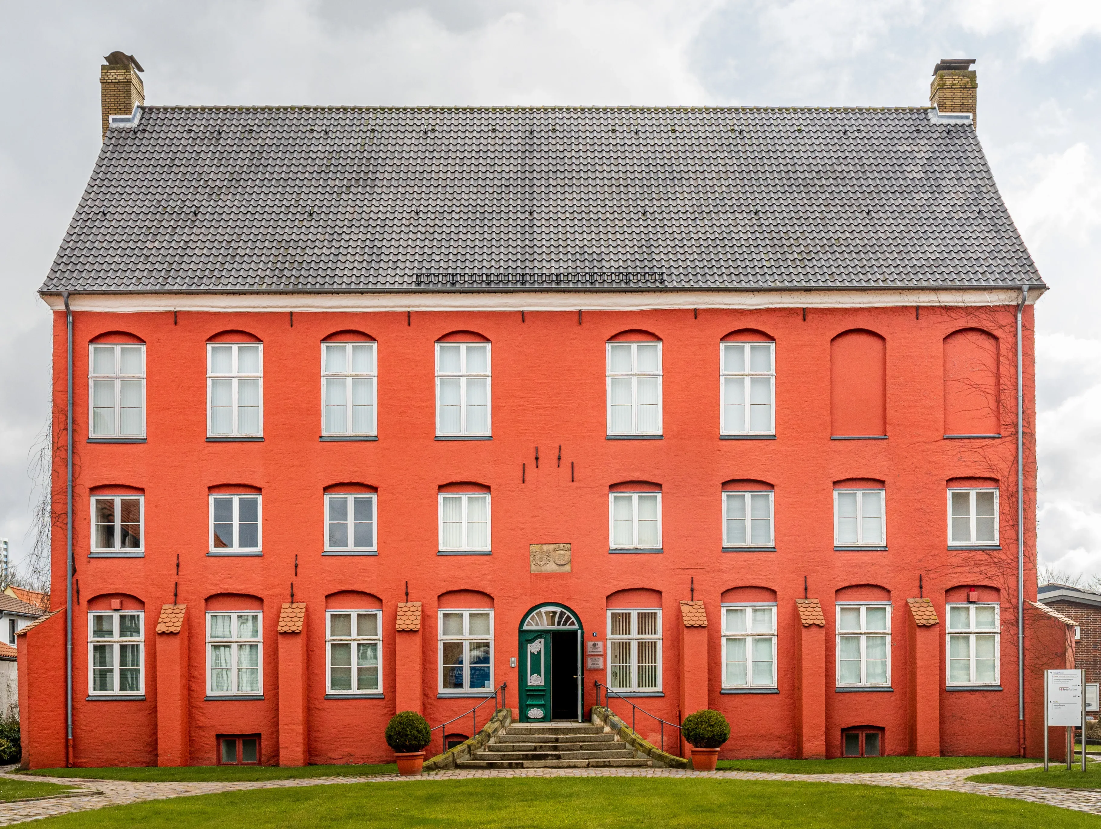 Historisches rotes Backsteingebäude mit Satteldach in Schleswig, bekannt als Günderrothscher Hof. Das Gebäude hat drei Stockwerke mit weißen Fenstern und einem dunkelgrünen Eingang in der Mitte, der über eine kleine Treppe erreichbar ist. Vor dem Eingang stehen zwei dekorative Pflanzen in Töpfen. Das Dach ist mit grauen Dachziegeln gedeckt, und im Vordergrund befindet sich eine gepflegte Rasenfläche.