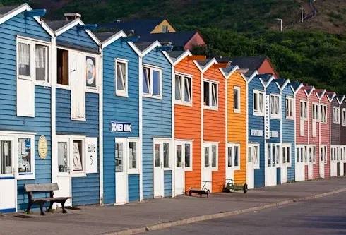 Bunte Reihenhäuser, die als 'Hummerbuden' bekannt sind, auf Helgoland, in kräftigen Farben wie Blau, Orange und Rot gestrichen, stehen entlang einer Promenade, mit Sitzbänken und einem ruhigen Hintergrund aus grünen Hügeln.