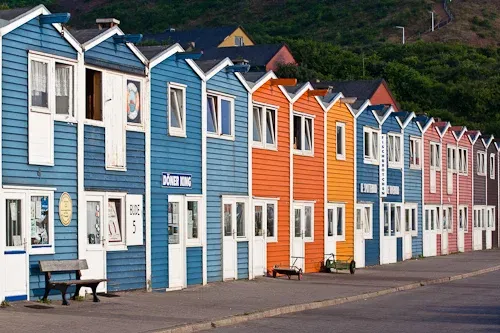 Bunte Reihenhäuser, die als 'Hummerbuden' bekannt sind, auf Helgoland, in kräftigen Farben wie Blau, Orange und Rot gestrichen, stehen entlang einer Promenade, mit Sitzbänken und einem ruhigen Hintergrund aus grünen Hügeln.