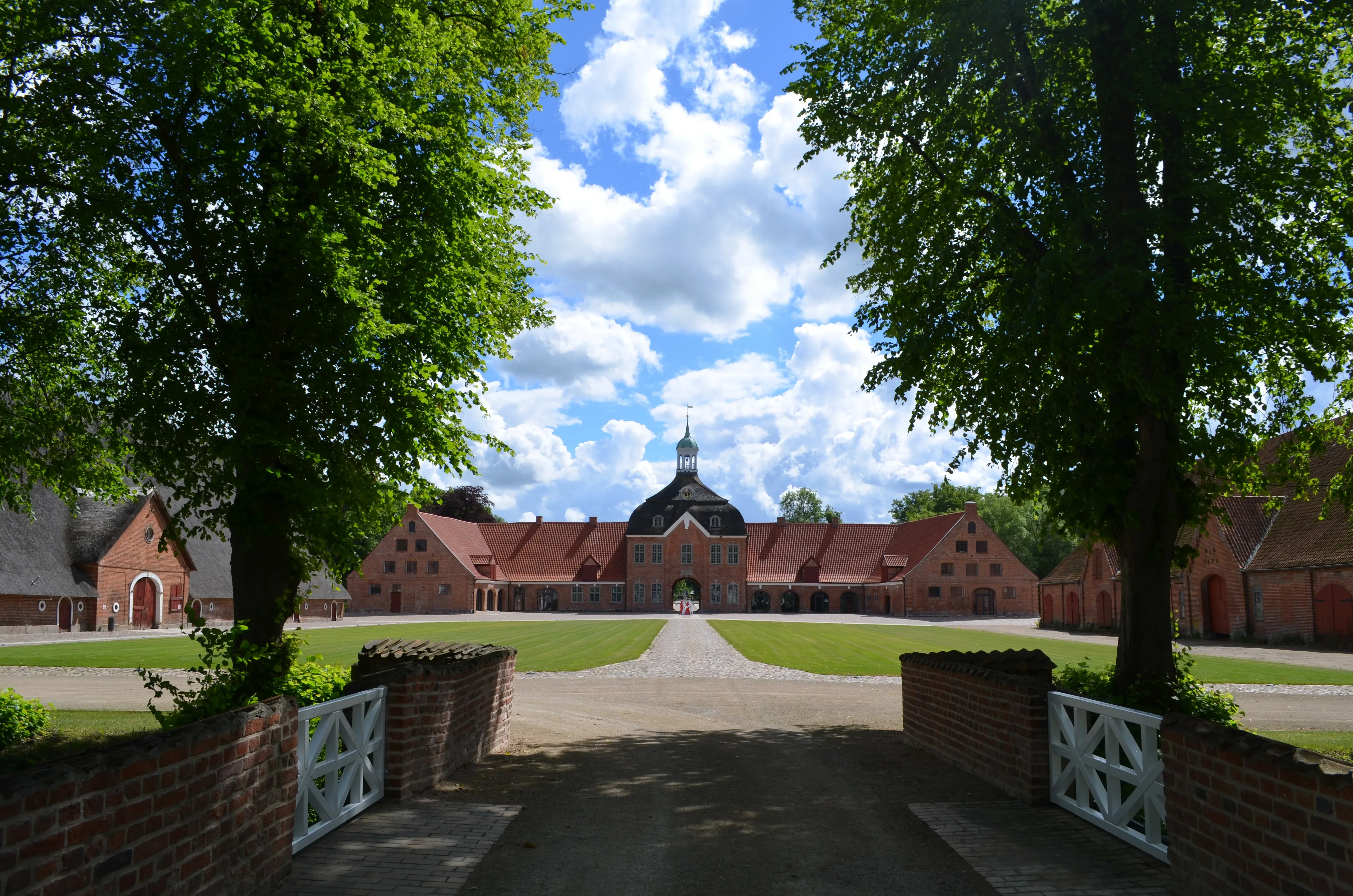 Blick durch eine von Bäumen gesäumte Allee auf das Torhaus von Gut Hasselburg, einem historischen Herrenhaus mit rotem Ziegeldach und symmetrischen Nebengebäuden, umgeben von gepflegten Rasenflächen und Backsteinmauern.