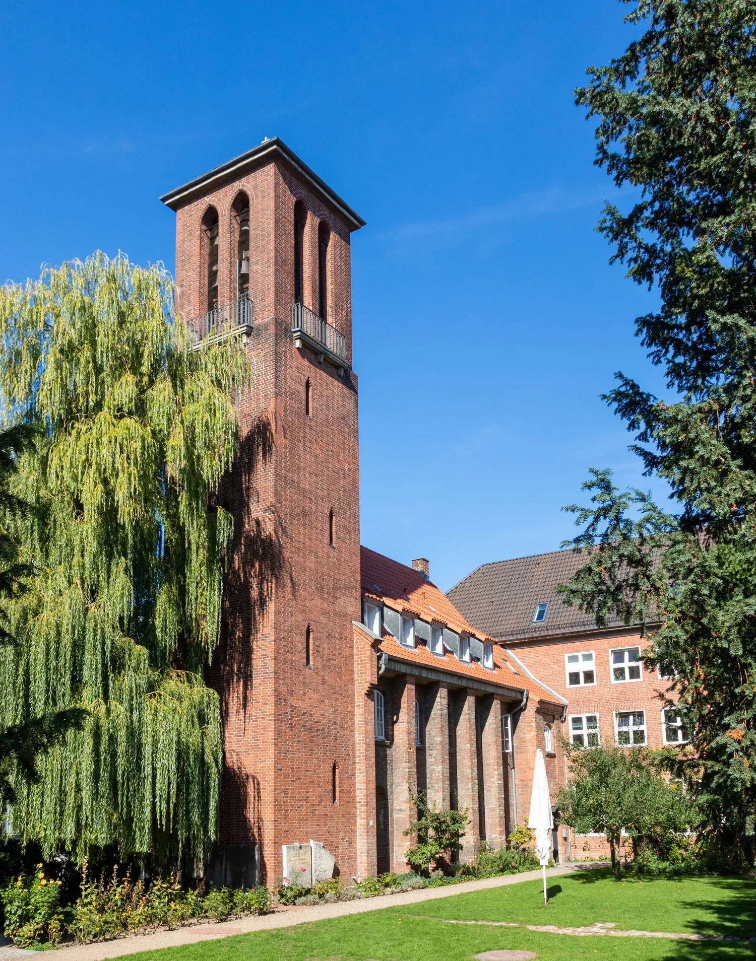 Ein roter Backsteinturm einer Kirche oder eines Klosters ragt in den blauen Himmel. Vor dem Gebäude steht eine große, grüne Trauerweide, die das Bild dominierend ergänzt. Die Architektur ist schlicht und weist klassische Backsteinelemente auf. Der Garten vor dem Gebäude ist gepflegt und von Bäumen umrahmt.