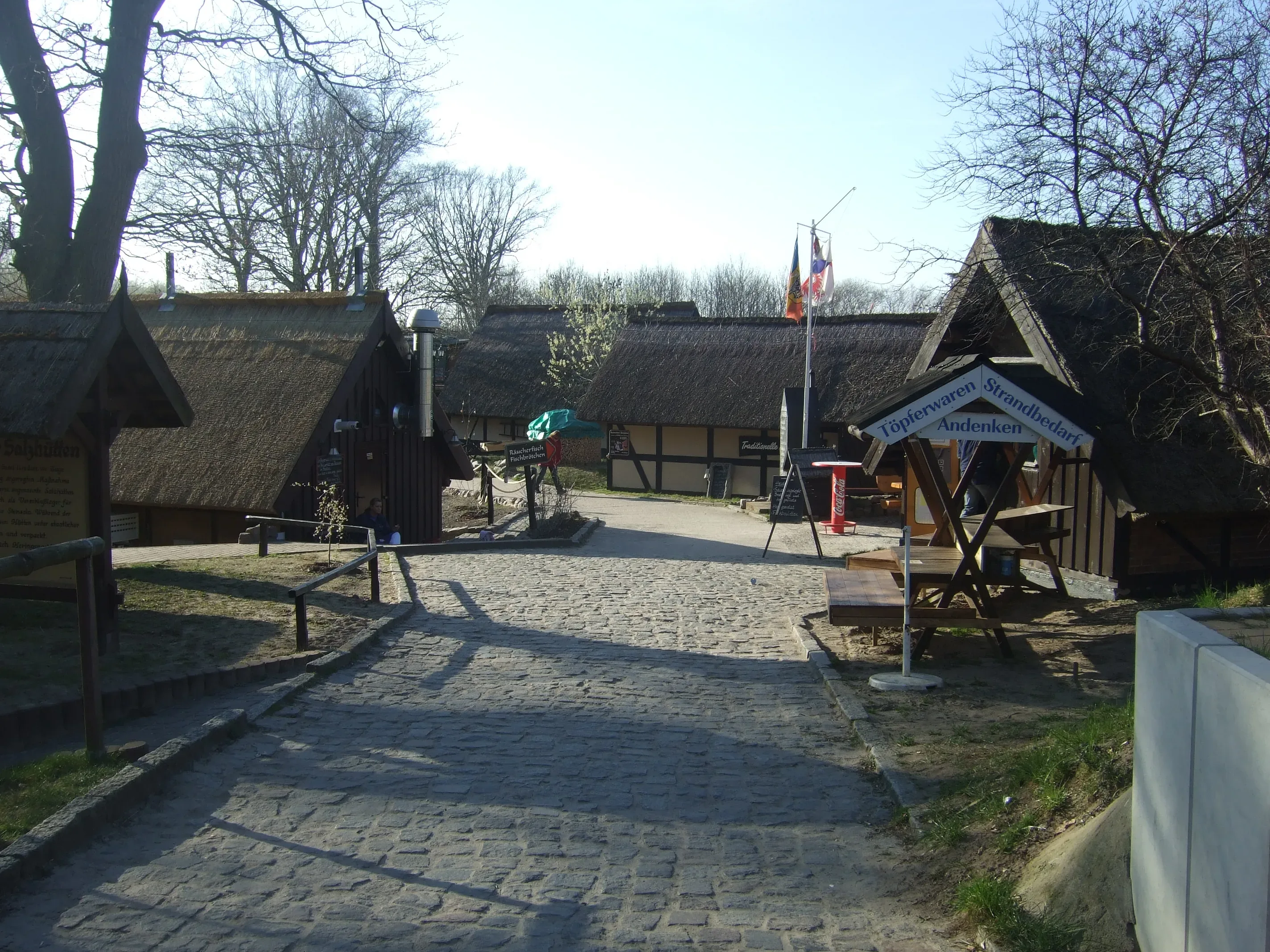 Blick auf die historischen Salzhütten in Koserow, die mit Strohdächern bedeckt sind und entlang eines gepflasterten Weges liegen, umgeben von Bäumen und kleinen Geschäften, die Souvenirs anbieten.