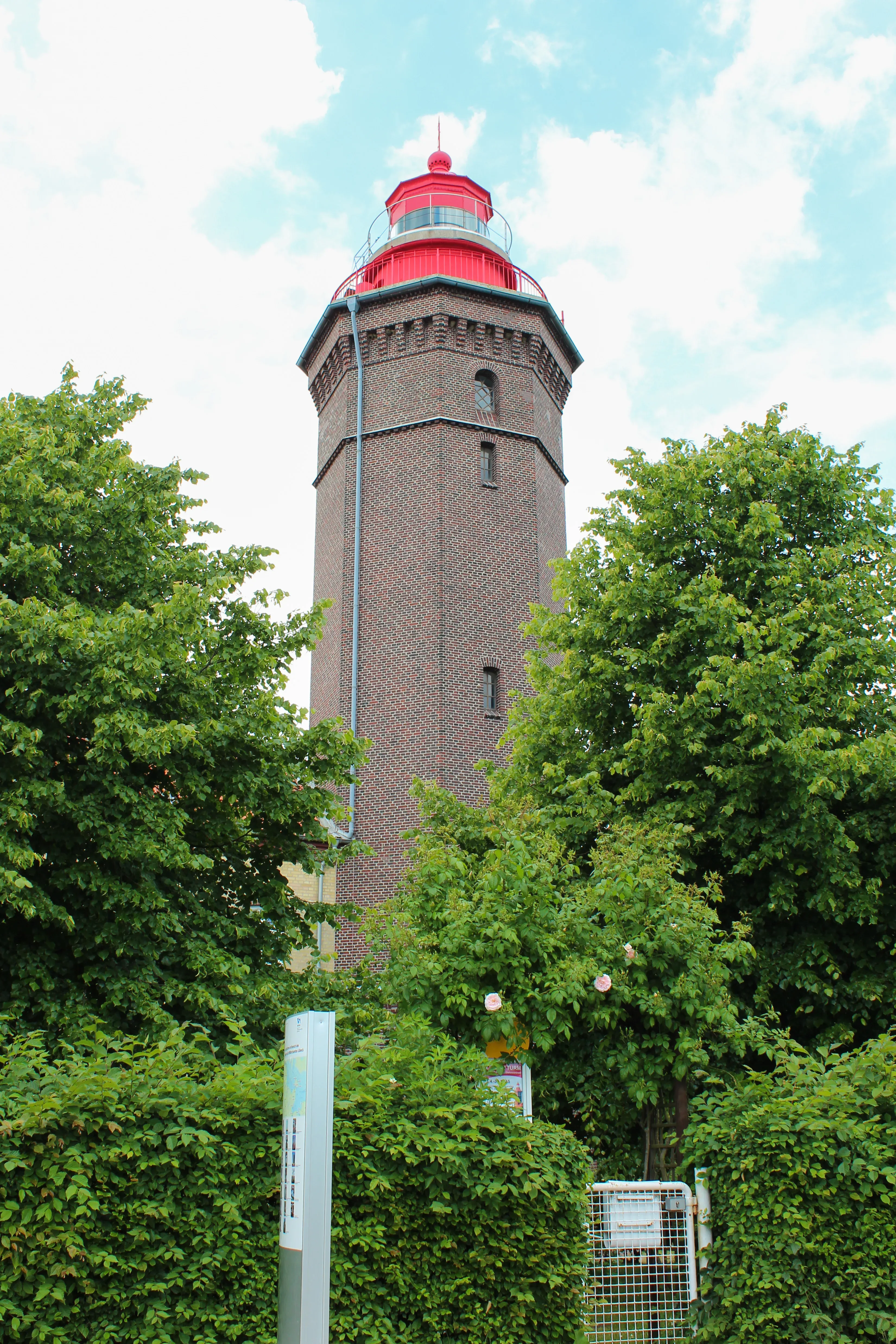 Ein rot gekrönter Backstein-Leuchtturm ragt zwischen grünen Bäumen in die Höhe, unter einem leicht bewölkten Himmel.