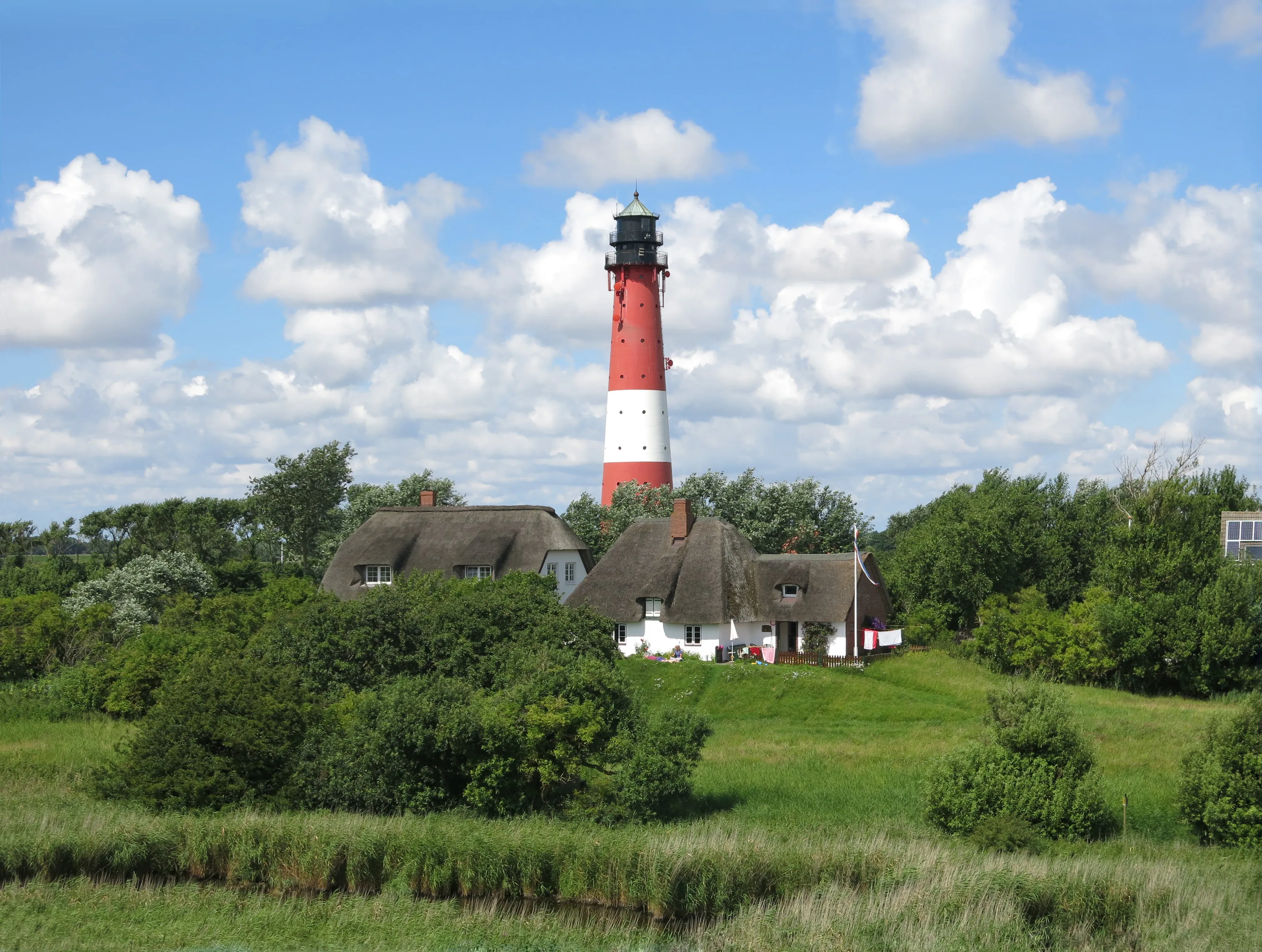 Der rot-weiße Leuchtturm auf Pellworm ragt hoch in den bewölkten Himmel, umgeben von Bäumen und einem malerischen Garten.