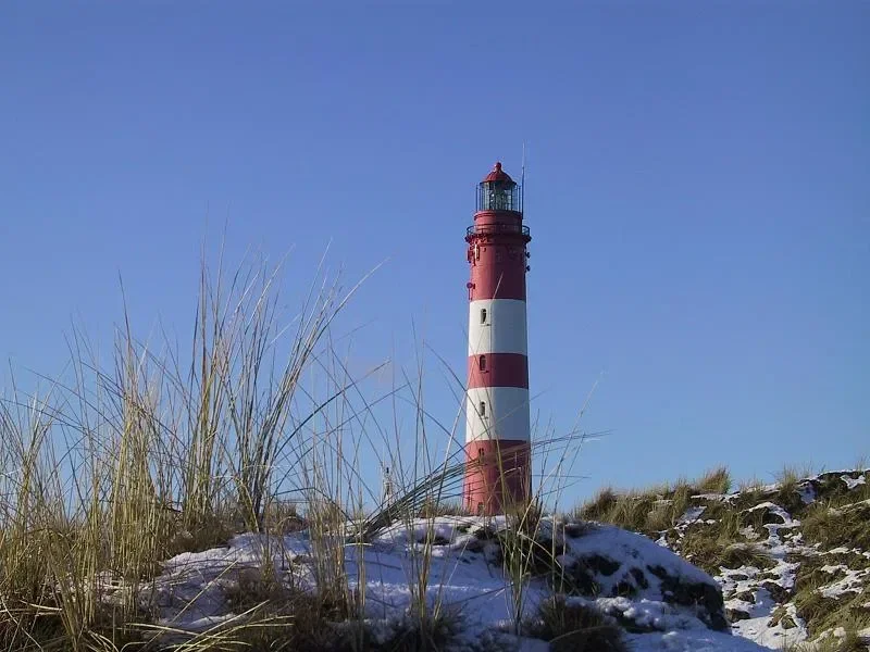 Der rot-weiße Leuchtturm auf Amrum ragt in den klaren Winterhimmel, während der Boden leicht mit Schnee bedeckt ist.