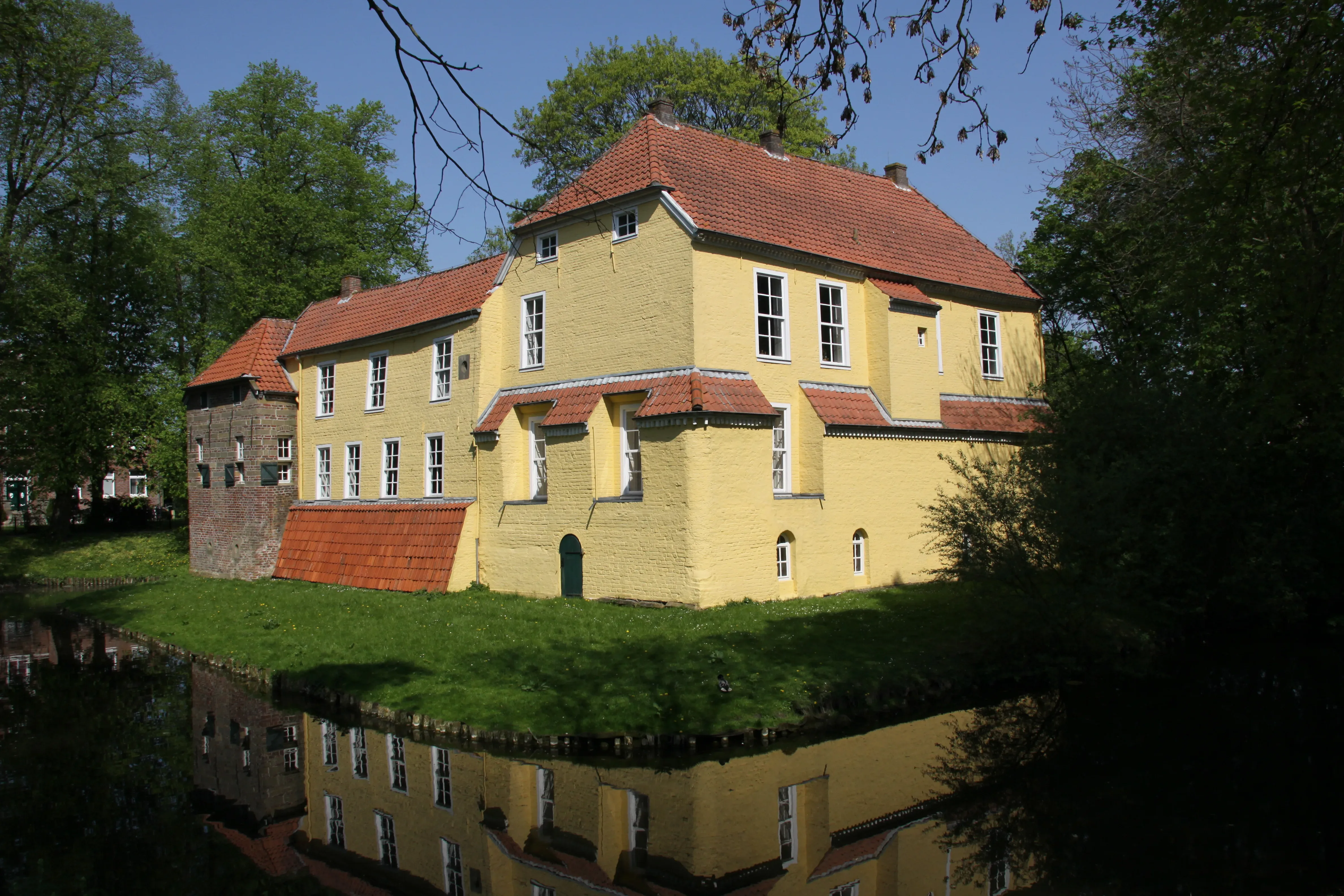 Ein gelbes Herrenhaus mit roten Ziegeldächern, umgeben von einem Wassergraben. Bäume und Sträucher säumen die Umgebung, und das Gebäude spiegelt sich im Wasser.