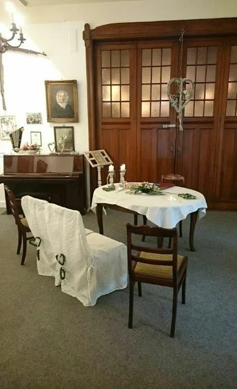 Ein kleiner, historischer Raum mit einer Hochzeitstafel, zwei weiß überzogenen Stühlen und dekorativem Herzschmuck, bereit für eine Trauung im Museum.