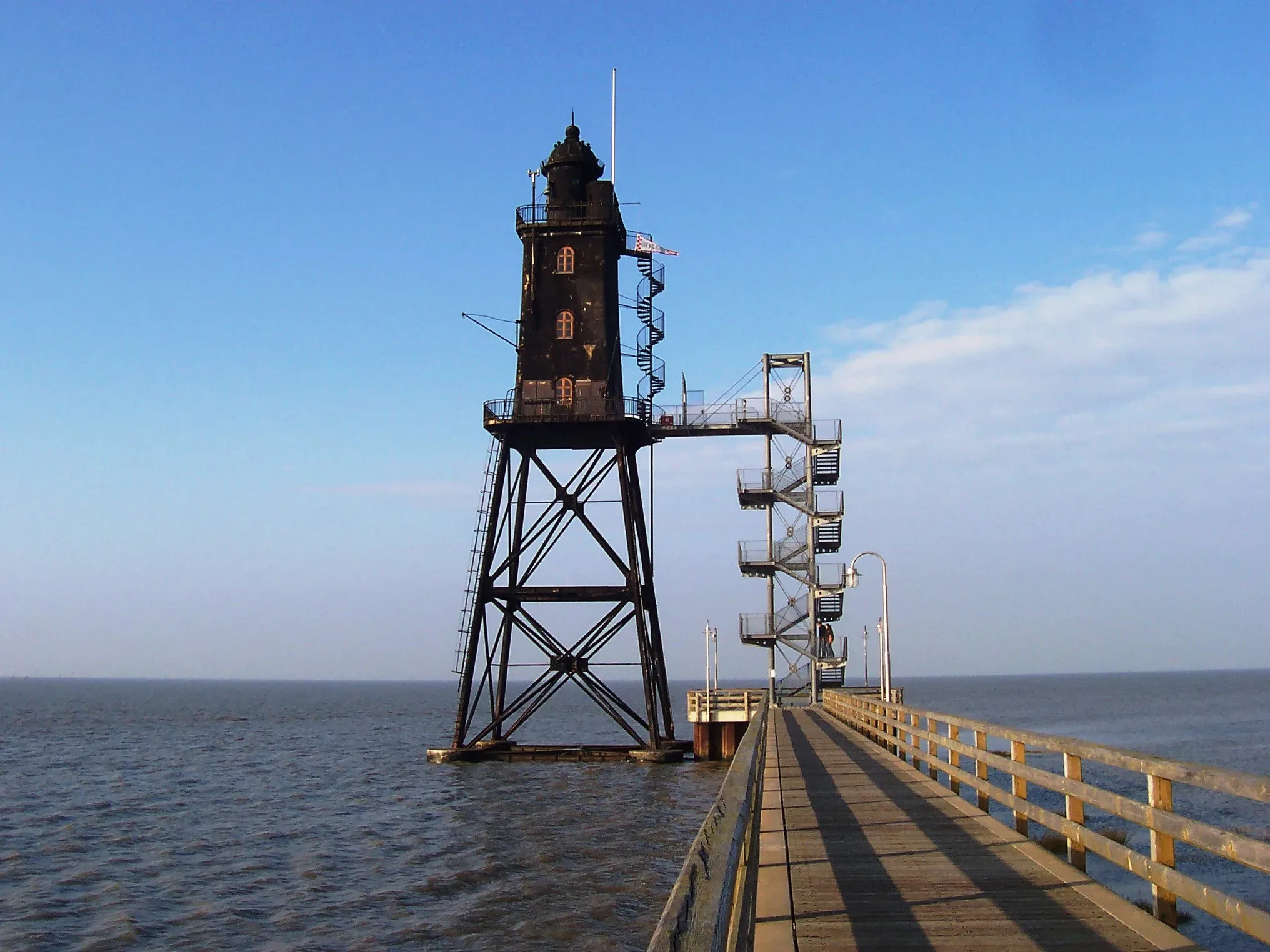Der Leuchtturm Obereversand mit einer markanten Metalltreppe, die von einem langen Holzsteg erreicht wird, ragt über dem ruhigen Meer auf.