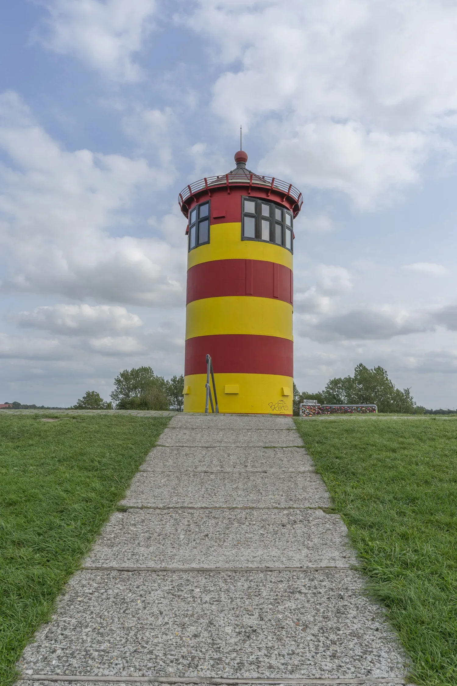 Der rot-gelb gestreifte Pilsumer Leuchtturm mit einem gepflasterten Weg, der zu seiner Tür führt, auf einem grasbewachsenen Hügel.
