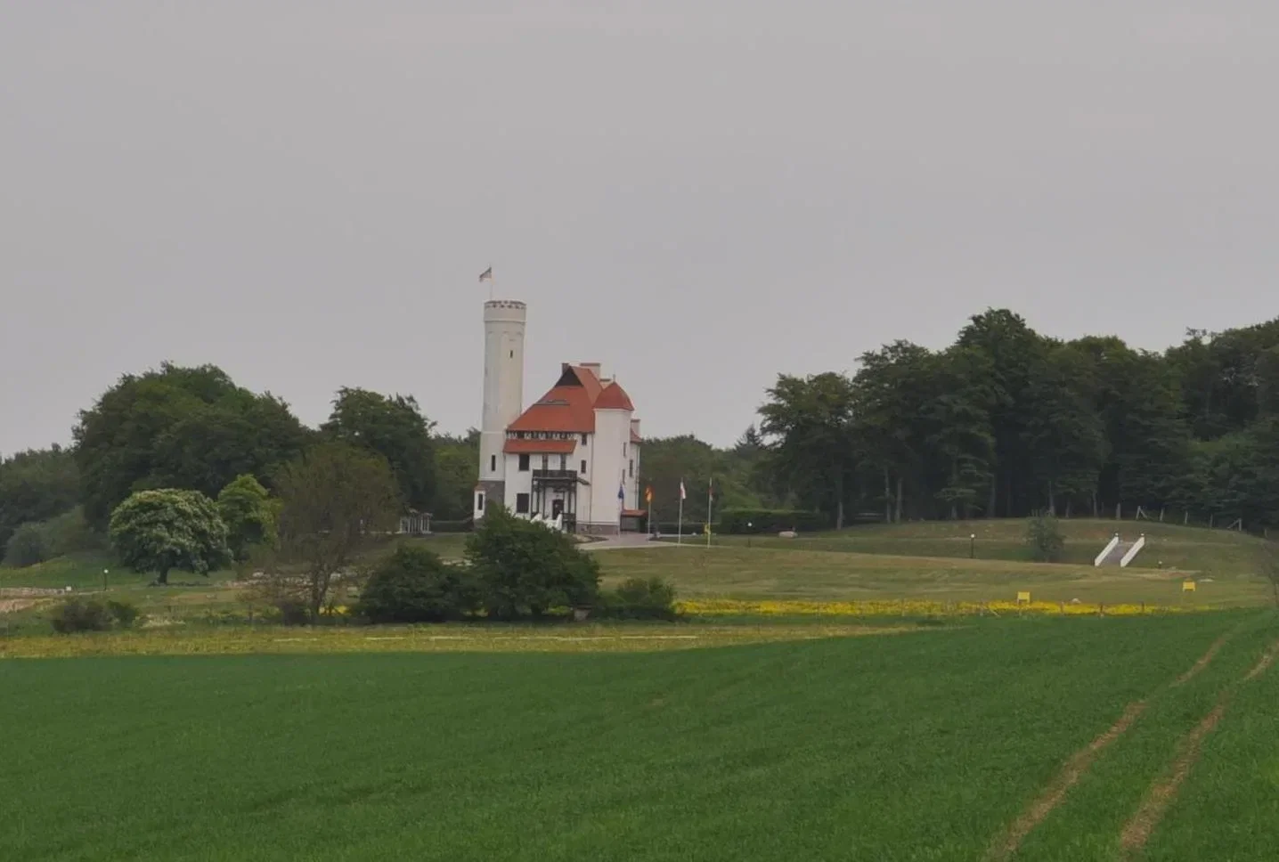 Weißes Schloss Ranzow mit Turm auf einem Hügel, umgeben von grünen Feldern und bewölktem Himmel.