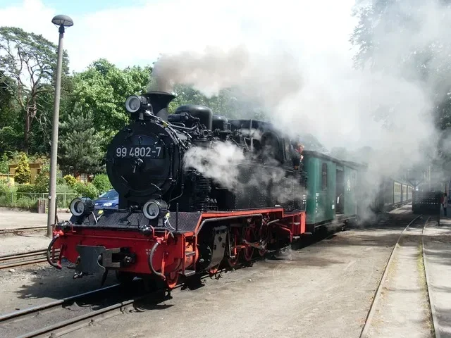 Die historische Dampflokomotive 'Rasender Roland' fährt auf einem Bahnhofsgleis, umgeben von dichten Bäumen, während Dampf aus ihrem Schornstein aufsteigt.