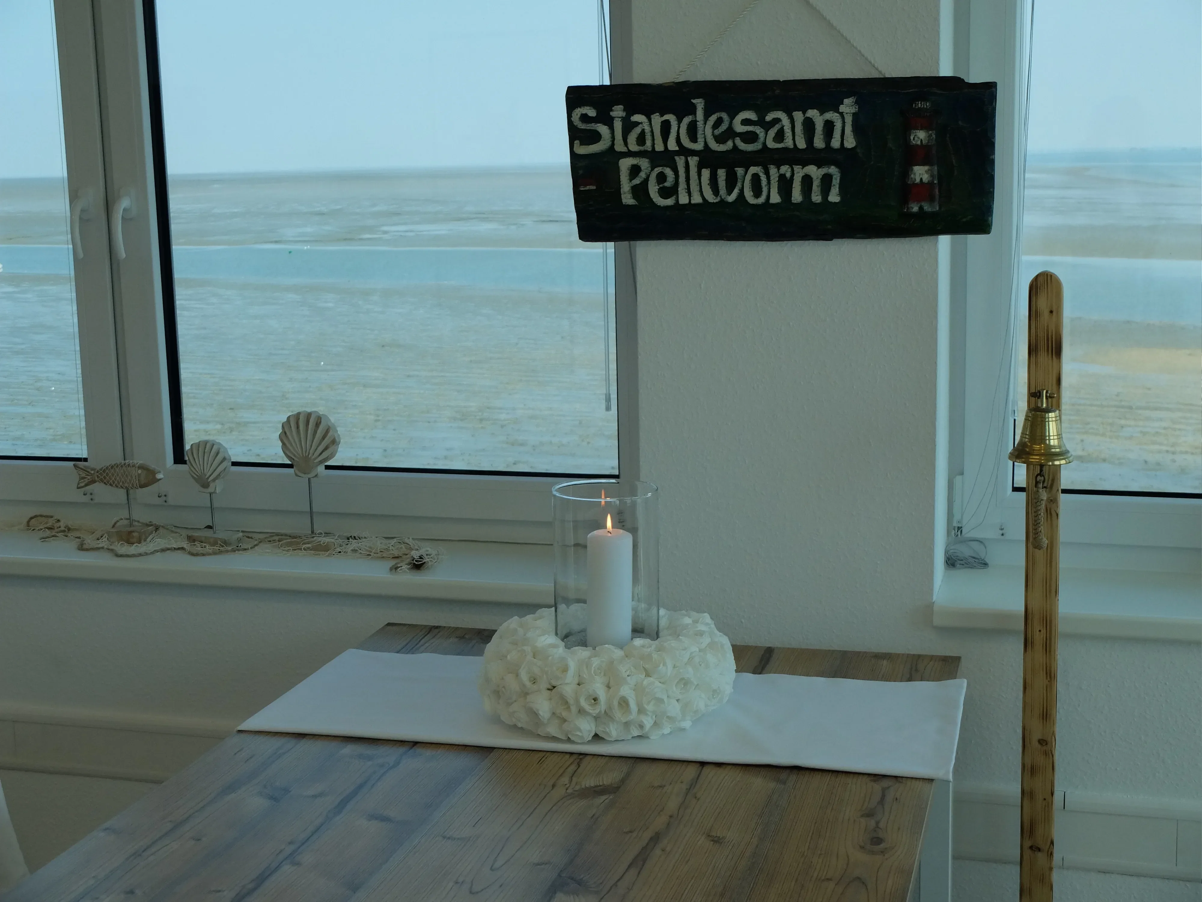 Ein Tisch im Standesamt Pellworm mit Blick auf die Nordsee. Der Tisch ist mit einer weißen Kerze und Muschel-Dekorationen geschmückt, während durch das Fenster das Wattenmeer sichtbar ist.