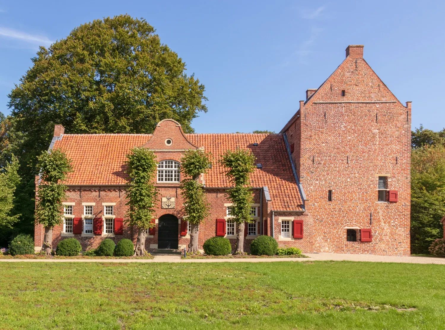 Historisches Steinhaus Bunderhee mit rotem Ziegeldach und leuchtend rotem Mauerwerk, umgeben von einem grünen Rasen und Bäumen im Hintergrund.