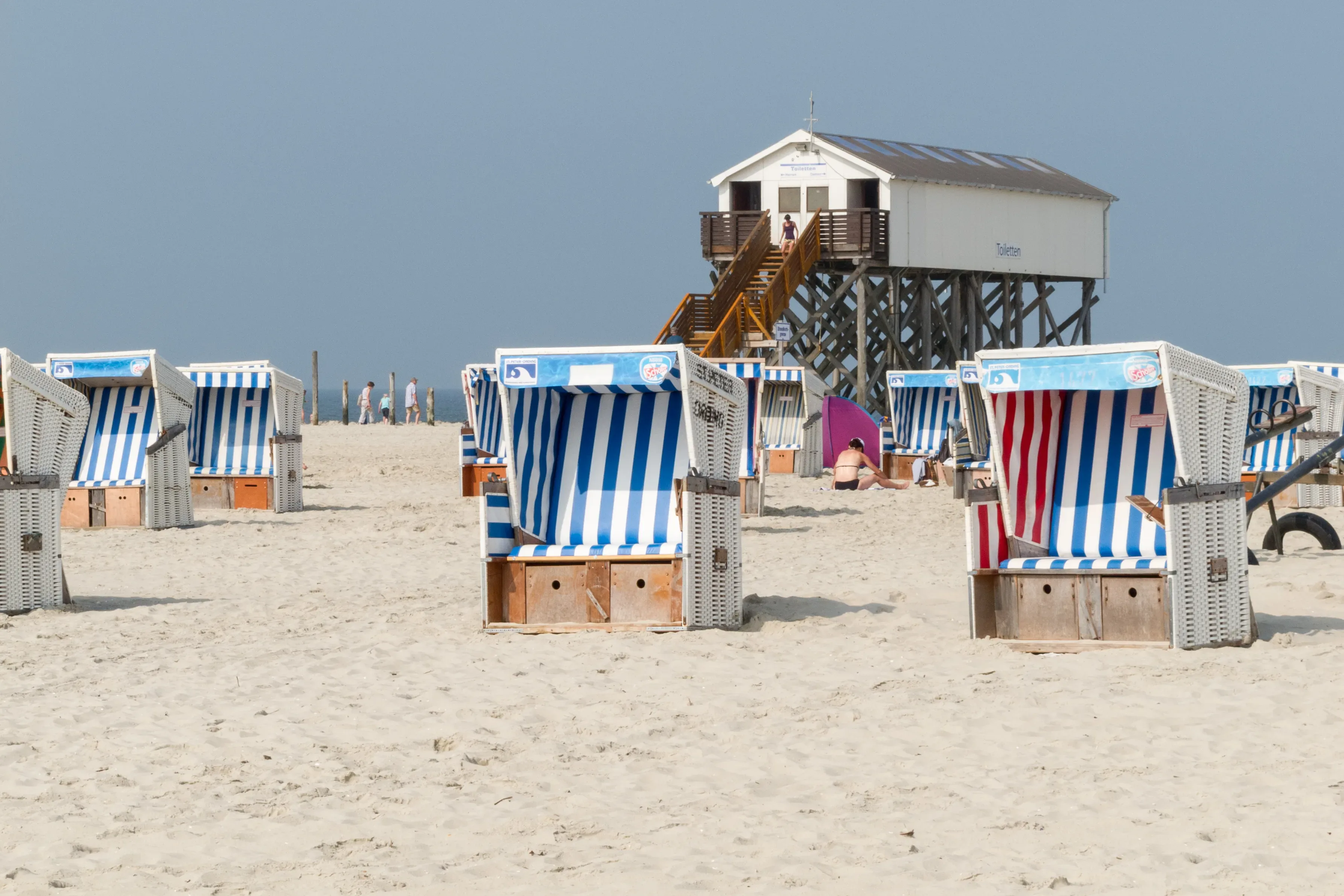 Blick auf den Strand von Sankt Peter-Ording, gesäumt mit blau-weiß gestreiften Strandkörben und einem Pfahlbau im Hintergrund, der auf Holzpfählen über dem Sand thront.