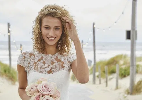 Eine lächelnde Braut mit lockigem Haar trägt ein weißes Spitzenkleid und hält einen Strauß rosafarbener Rosen. Im Hintergrund ist ein Strand mit Lichterketten zu sehen.