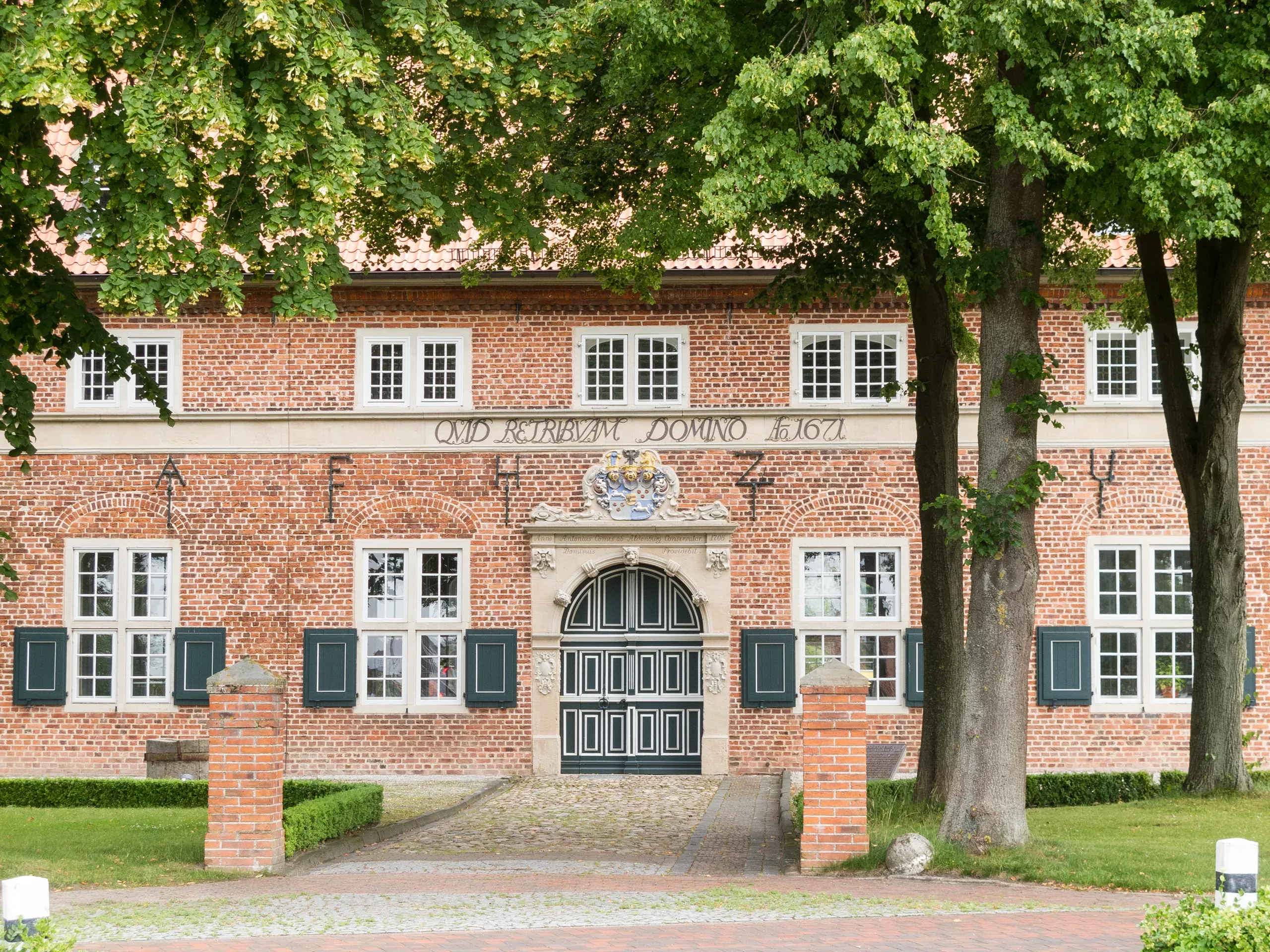 Historisches Waisenhaus in Varel mit Backsteinfassade, großen grünen Türen und Fenstern, umgeben von Bäumen und einer gepflegten Gartenanlage.