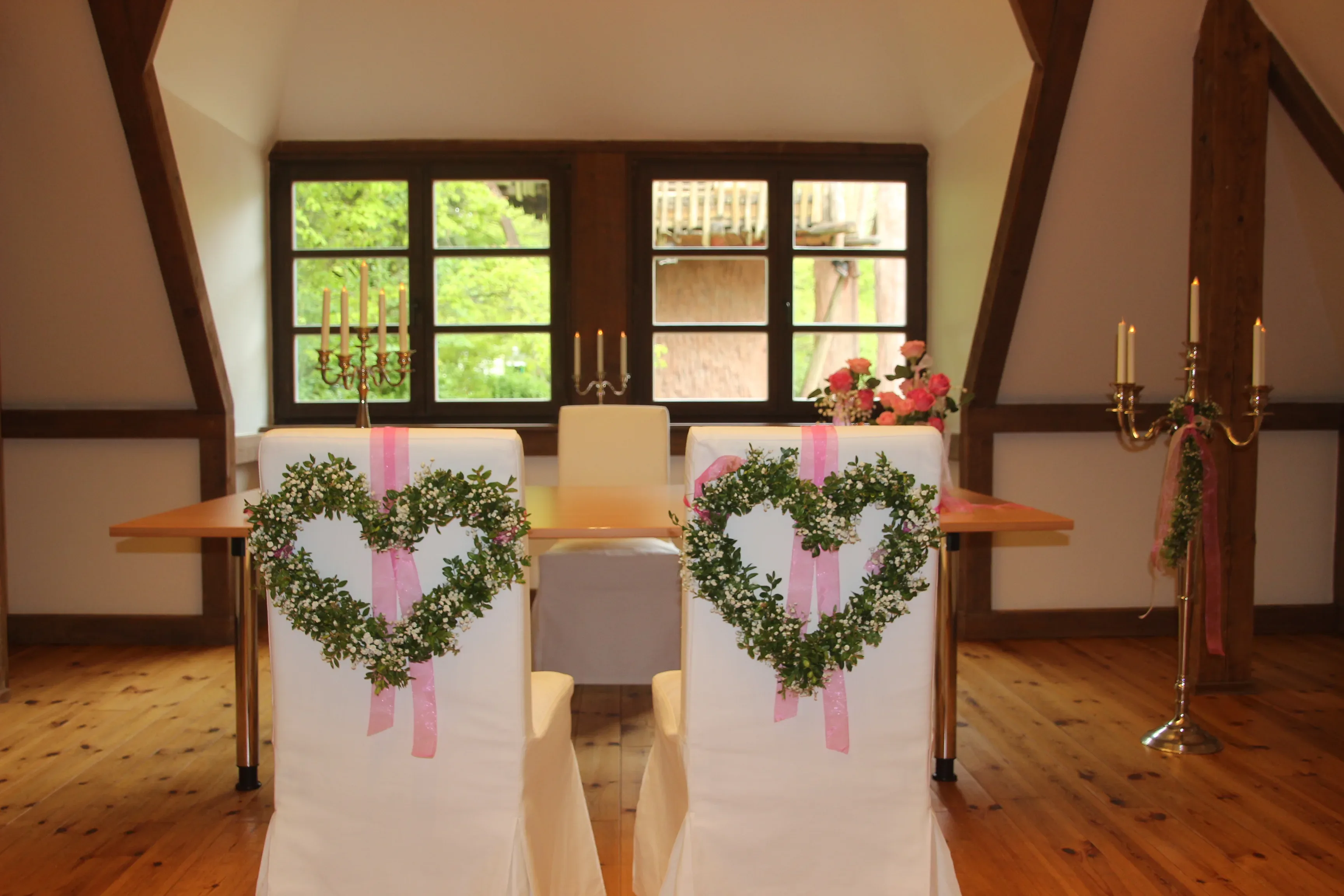 Ein festlich geschmückter Trauungsraum im Hansapark, mit weißen Stühlen, die mit herzförmigen Blumengestecken und rosa Schleifen dekoriert sind, und einem Fenster mit Blick ins Grüne.
