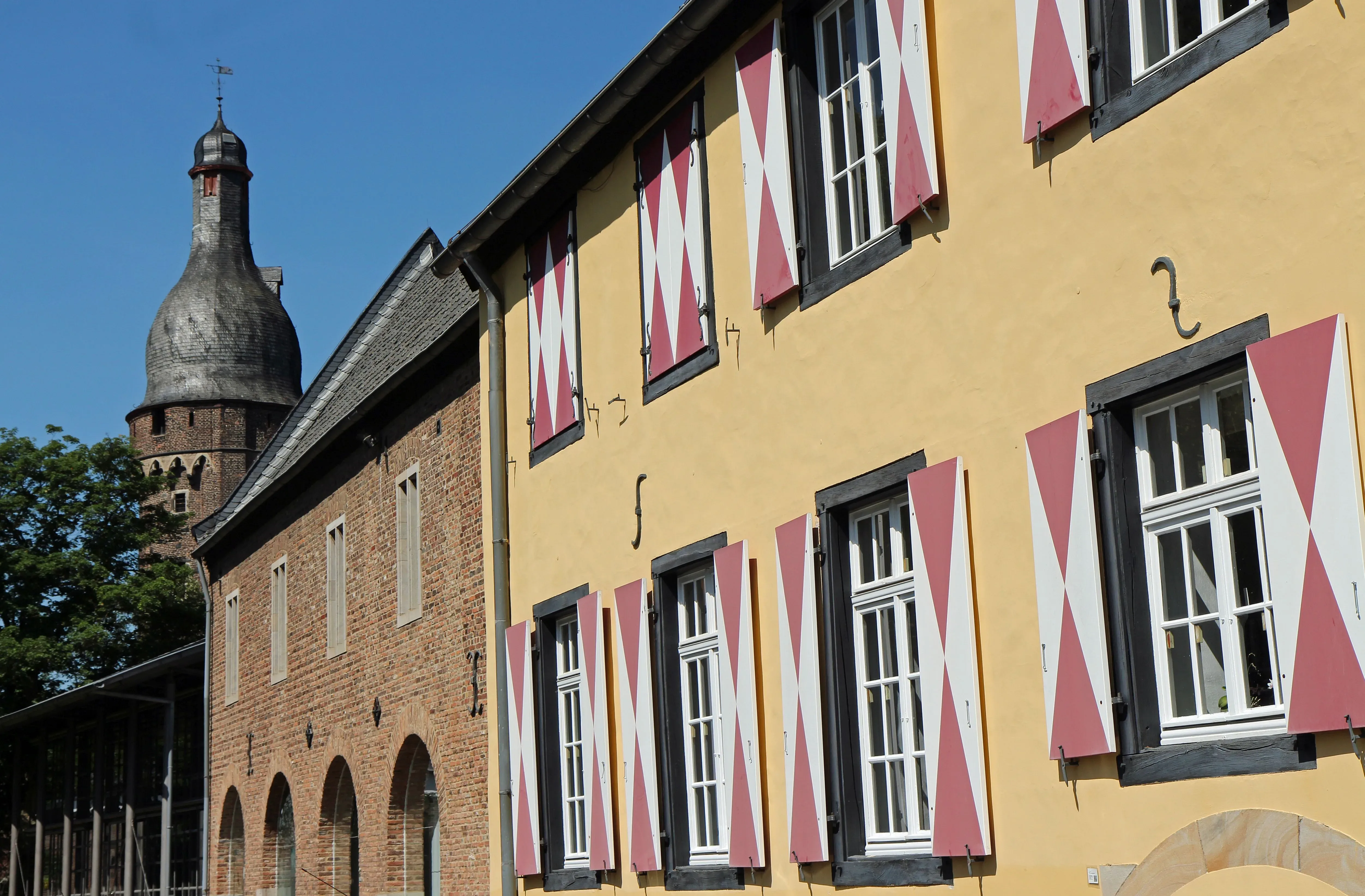 Schlossähnliches Gebäude mit markantem Turm und rot-weiß gestreiften Fensterläden an einem sonnigen Tag.