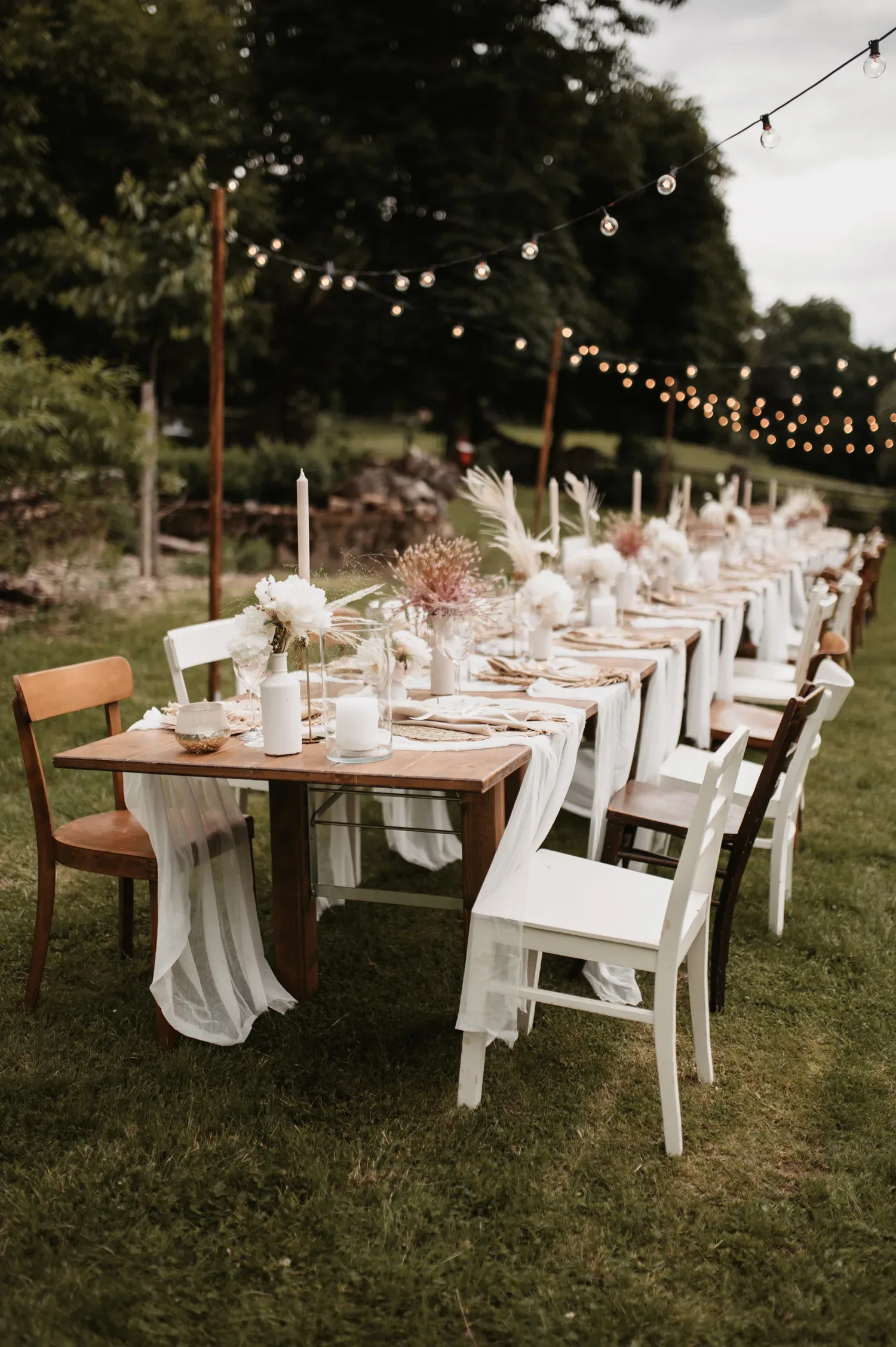 Eine lange Hochzeitstafel im Freien, dekoriert mit weißen Blumen, Kerzen und Boho-Elementen. Verschiedene Stühle und Tischläufer in Weiß sorgen für eine entspannte, rustikale Atmosphäre, während Lichterketten über dem Tisch die Szene beleuchten.