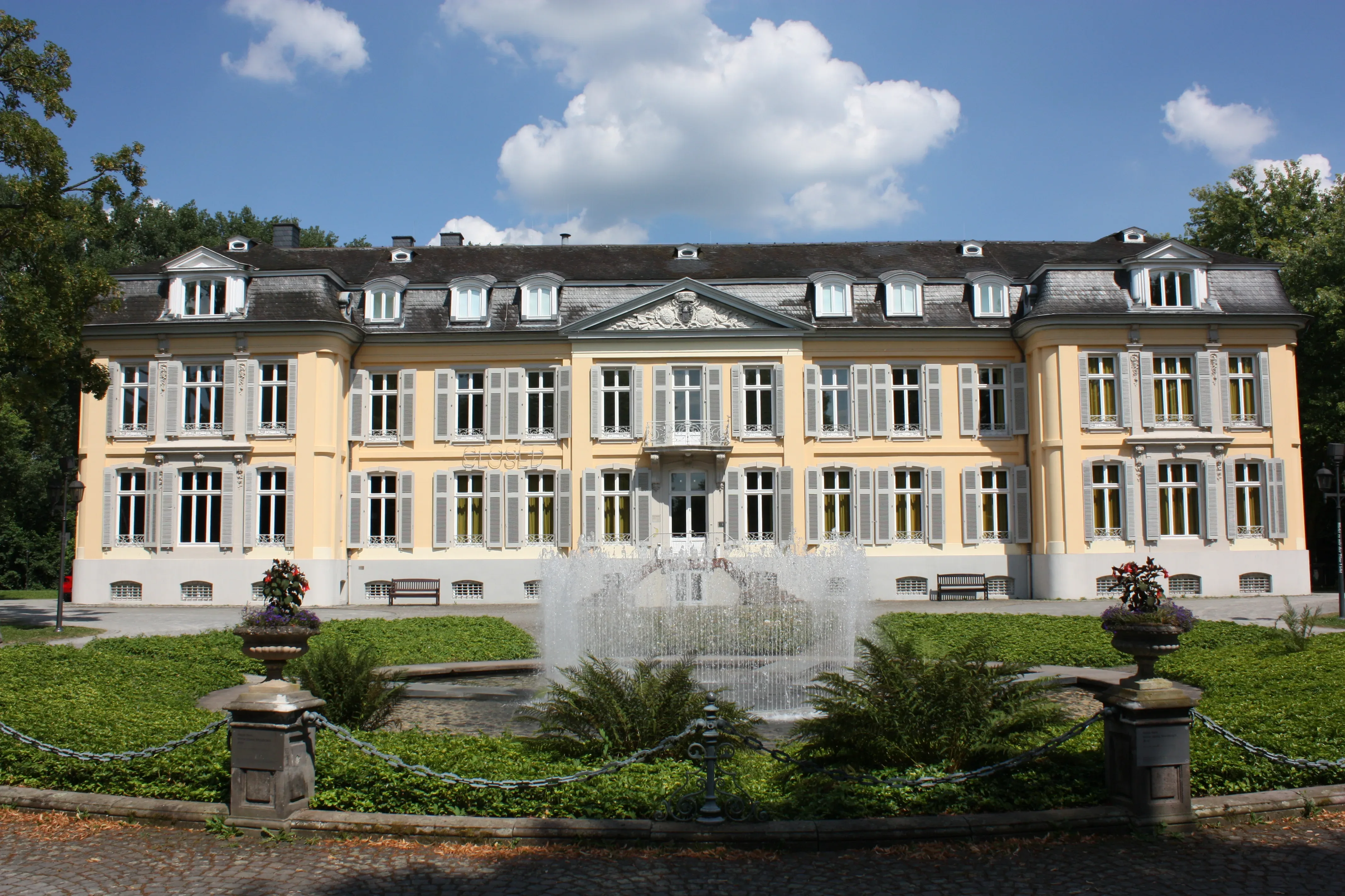 Ein barockes Schloss mit gepflegten Gärten und einem eindrucksvollen Springbrunnen, umgeben von blauen Sommerwolken.
