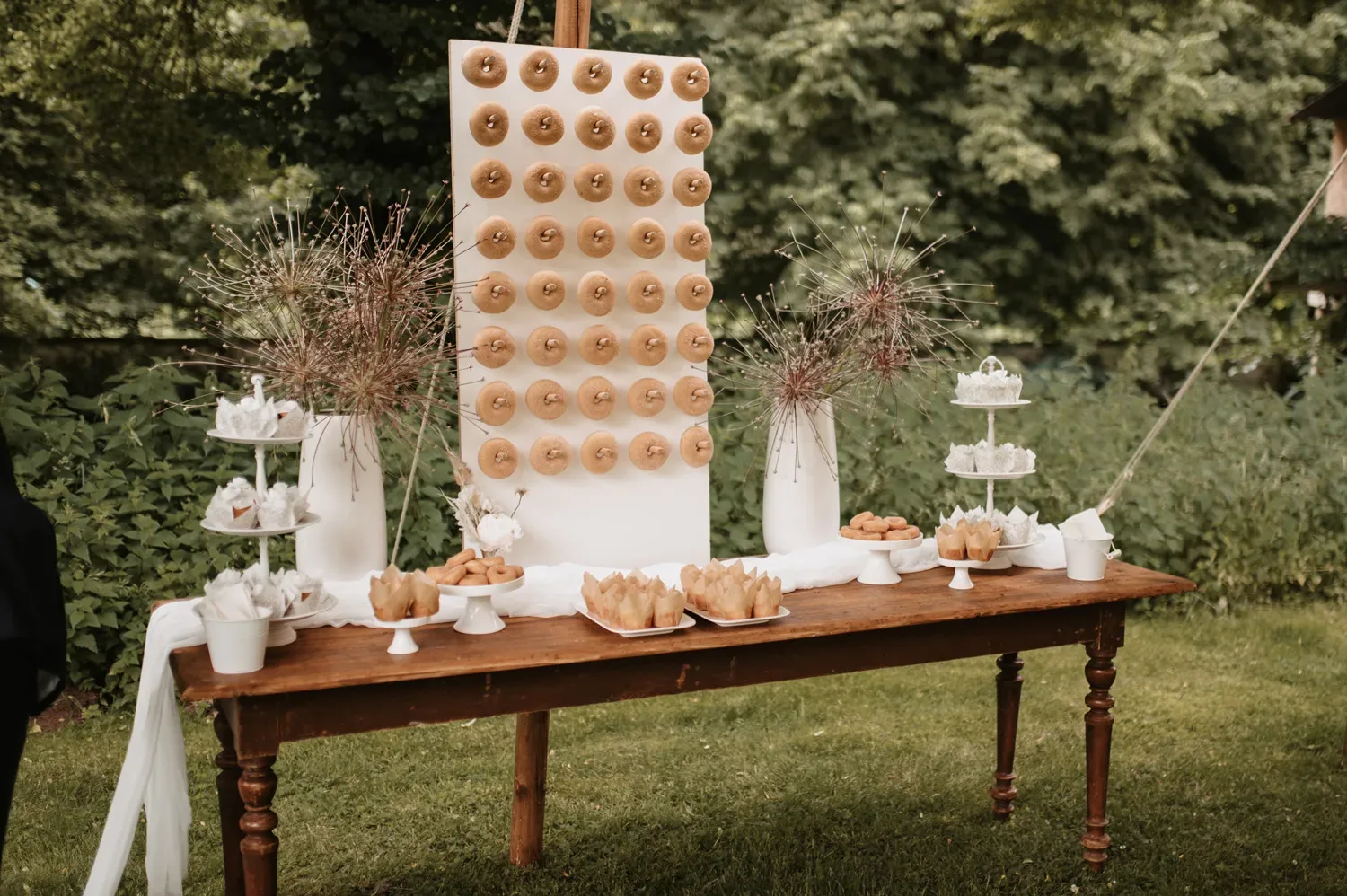 Ein rustikaler Hochzeitstisch im Freien, dekoriert mit einer Donut-Wand, auf der mehrere Reihen von Donuts auf Holzstangen hängen. Der Tisch ist mit weißen Vasen, Trockenblumen und verschiedenen süßen Leckereien geschmückt.