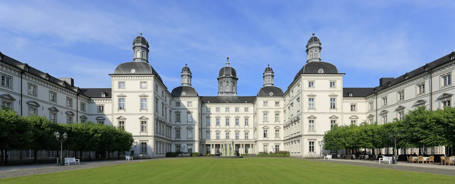Außenansicht von Schloss Bensberg mit imposanter Fassade und symmetrischer Architektur, unter strahlend blauem Himmel und umgeben von gepflegtem Rasen.