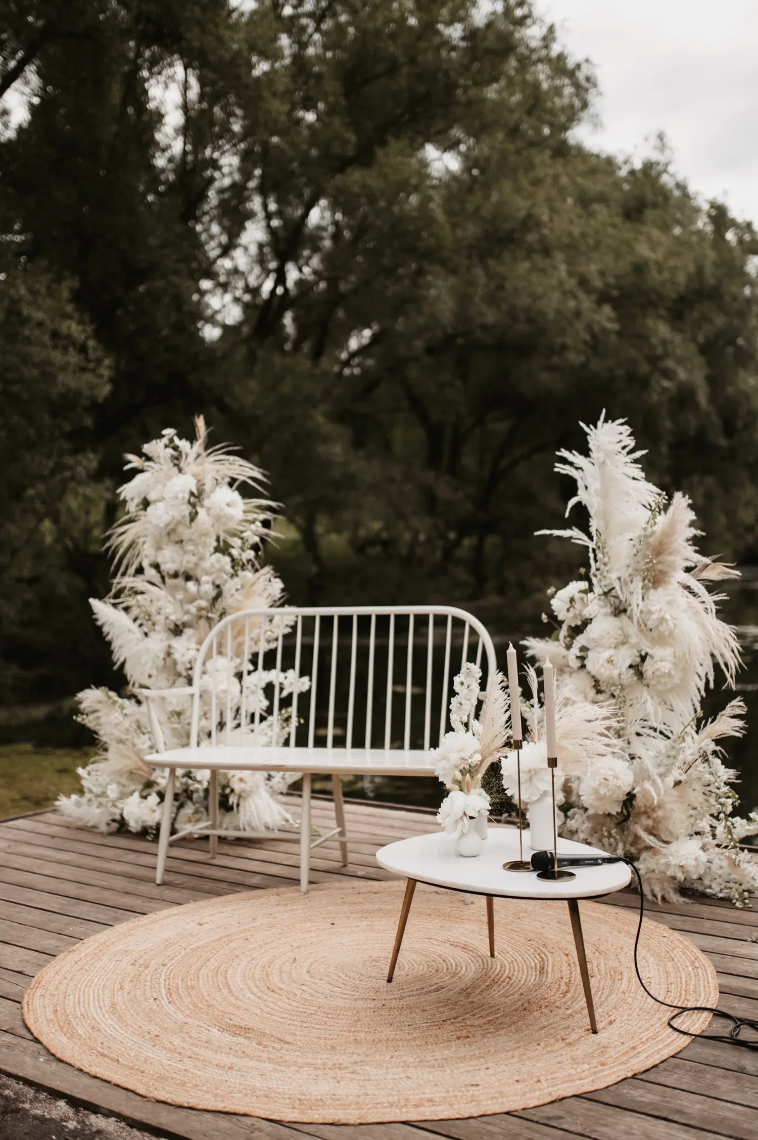 Eine elegante, weiße Bank steht auf einem Holzpodest, umgeben von kunstvoll arrangierten Pampasgras- und Blumengestecken. Ein runder Teppich und ein kleiner Tisch mit Blumen und Kerzen verleihen der Szene einen intimen, rustikalen Charme.