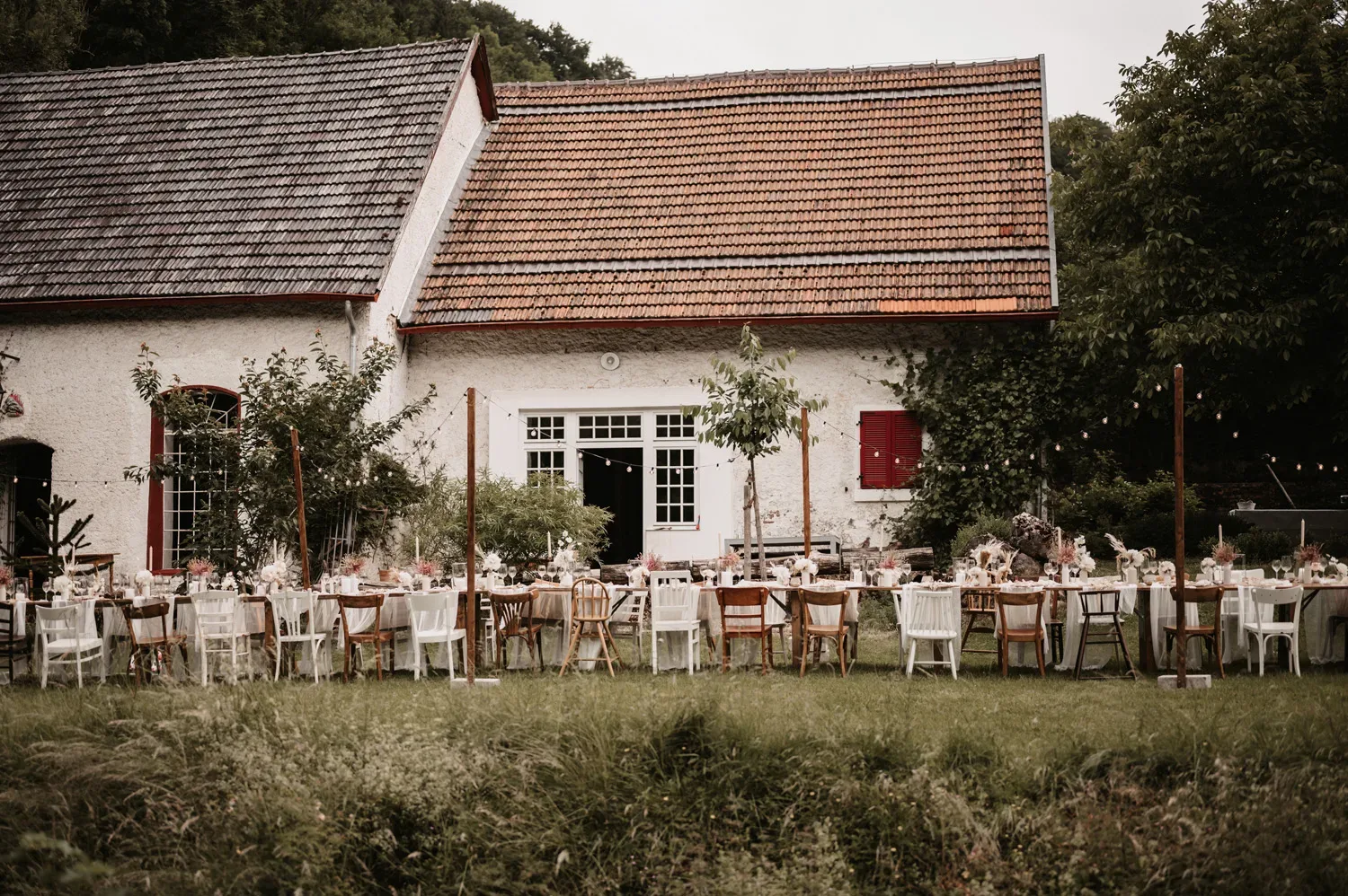 Ein idyllischer Außenbereich mit einer langen Tafel für eine Hochzeit, umgeben von einer rustikalen Scheune mit roten Fensterläden. Verschiedene Stühle und Lichterketten schaffen eine festliche und gemütliche Atmosphäre für die Gäste.