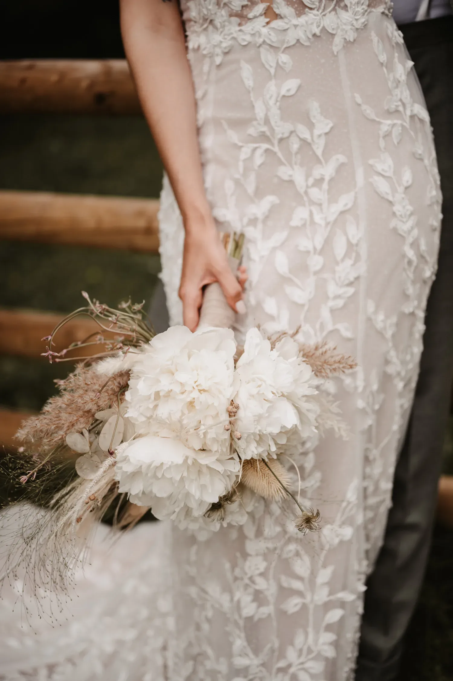 Nahaufnahme eines Brautstraußes mit weißen Pfingstrosen, Pampasgras und getrockneten Gräsern, gehalten von einer Braut in einem detailreichen, floralen Spitzenkleid. Die natürliche und rustikale Zusammenstellung des Straußes ergänzt das Boho-Thema der Hochzeit.