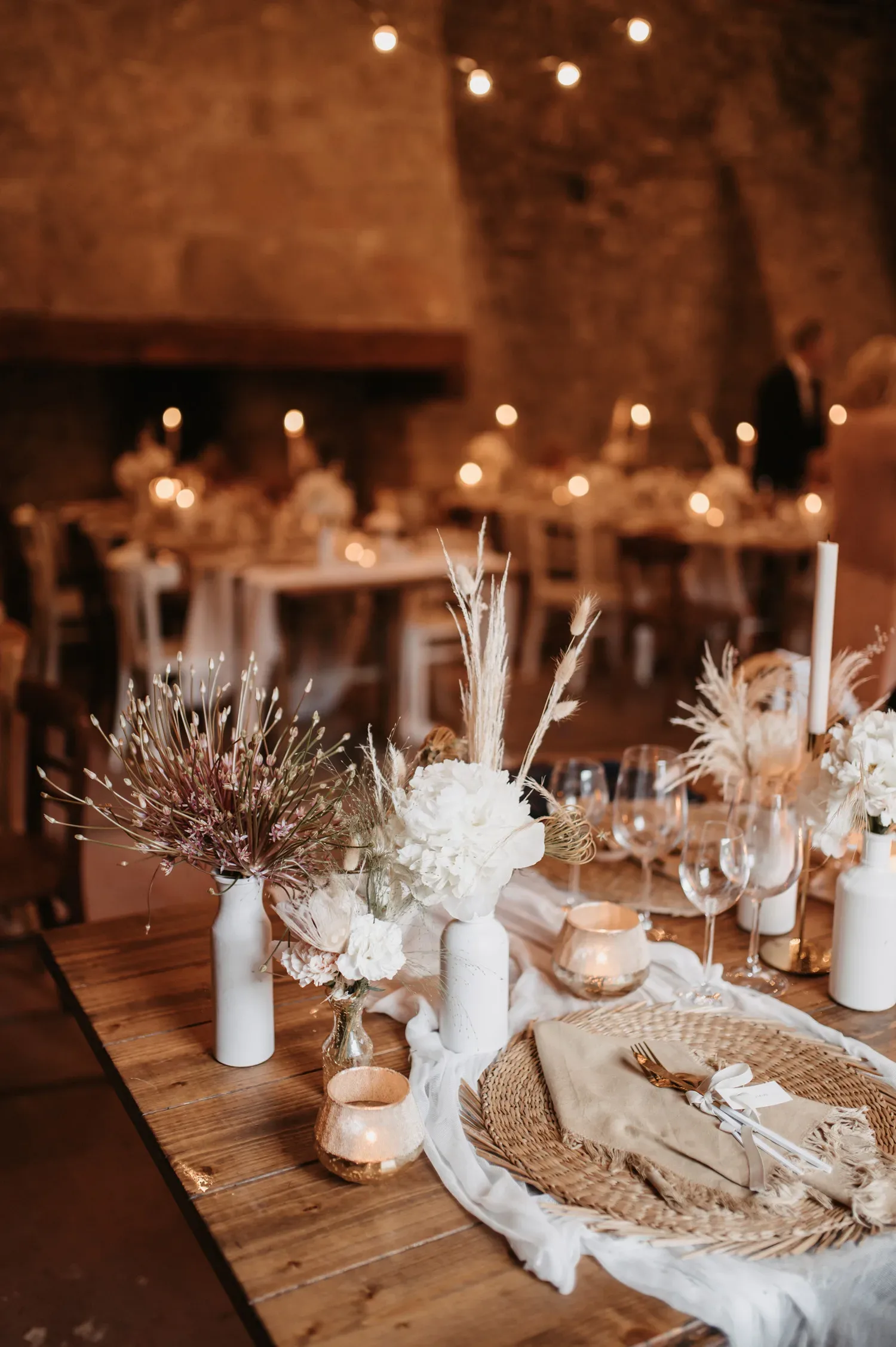 Ein festlich gedeckter Hochzeitstisch in einem rustikalen Innenraum, dekoriert mit Boho-Elementen wie getrocknetem Pampasgras und weißen Blumen. Kerzen und goldene Details sorgen für eine warme, romantische Atmosphäre.