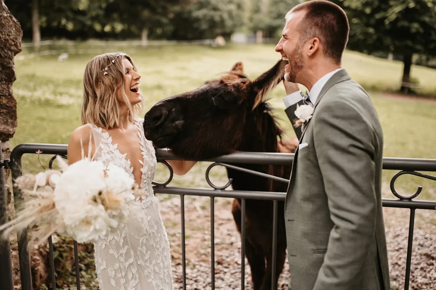 Ein lachendes Brautpaar steht an einem Zaun auf einer Wiese. Die Braut, in einem eleganten Spitzenkleid, hält einen Strauß weißer Blumen. Neben ihr steckt ein Esel seinen Kopf durch den Zaun, während der Bräutigam, der einen grauen Anzug trägt, ebenfalls lacht. Die Szene strahlt eine fröhliche, ungezwungene Atmosphäre in ländlicher Umgebung aus.
