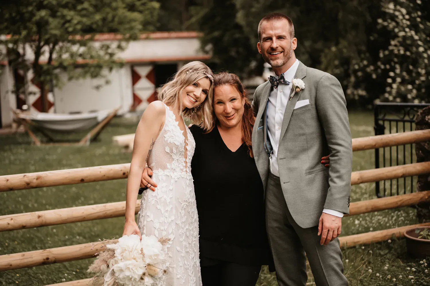 Das Brautpaar steht lächelnd mit einer dritten Person, möglicherweise der Hochzeitsplanerin, vor einem Zaun in einer grünen, ländlichen Umgebung. Die Braut trägt ein wunderschönes Spitzenkleid und hält einen Strauß weißer Blumen, während der Bräutigam einen grauen Anzug und eine Fliege trägt. Alle wirken entspannt und glücklich.