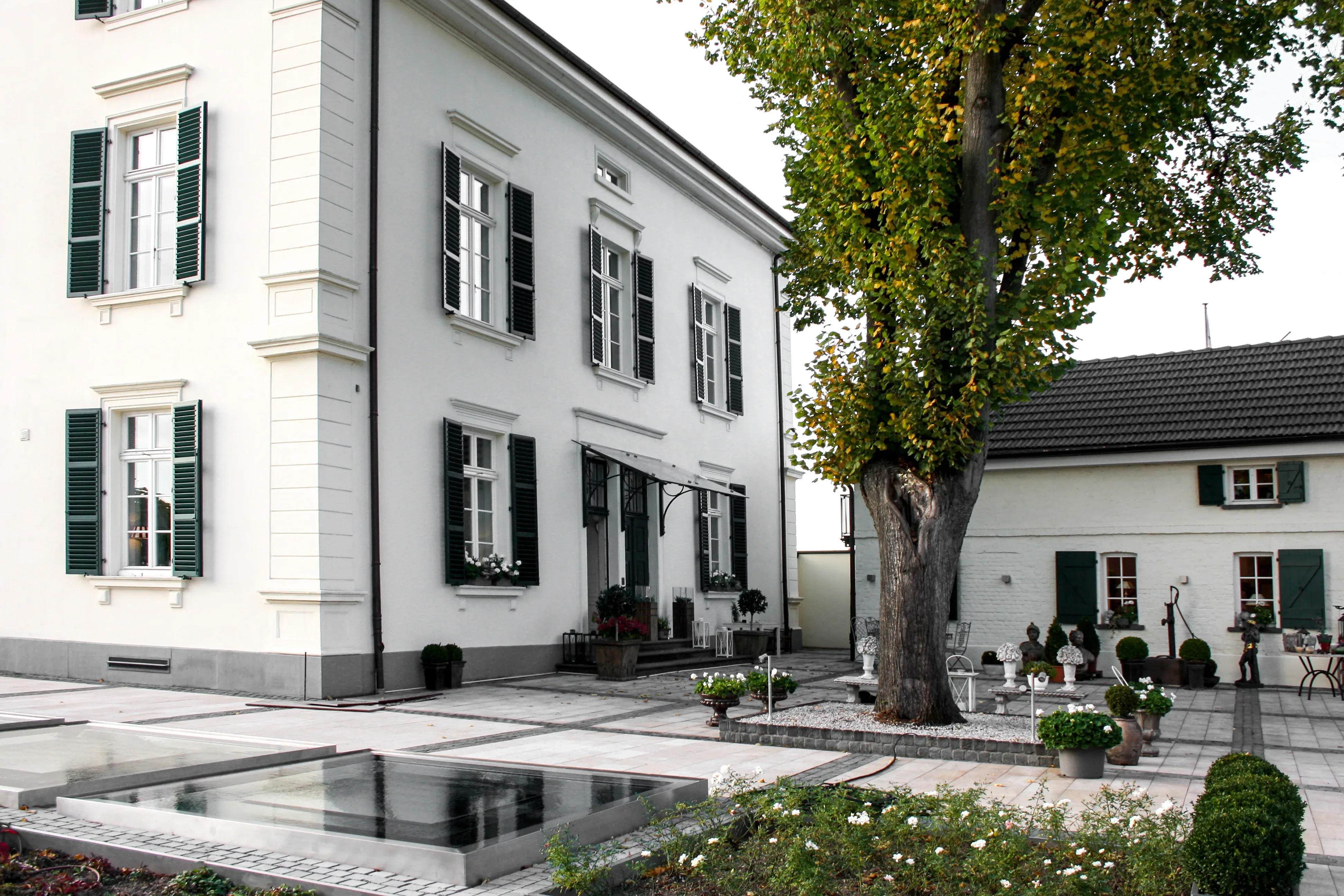 Außenansicht eines weiß gestrichenen Herrenhauses mit grünen Fensterläden und einem großen Baum im Hof. Der moderne, gepflasterte Vorplatz umfasst Wasserbecken und Sitzgelegenheiten.