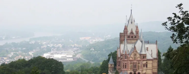 Weitläufiger Blick auf das Schloss Drachenburg in Königswinter, das majestätisch auf einem Hügel thront, umgeben von grünen Bäumen und mit einer leicht nebligen Stadtlandschaft im Hintergrund.